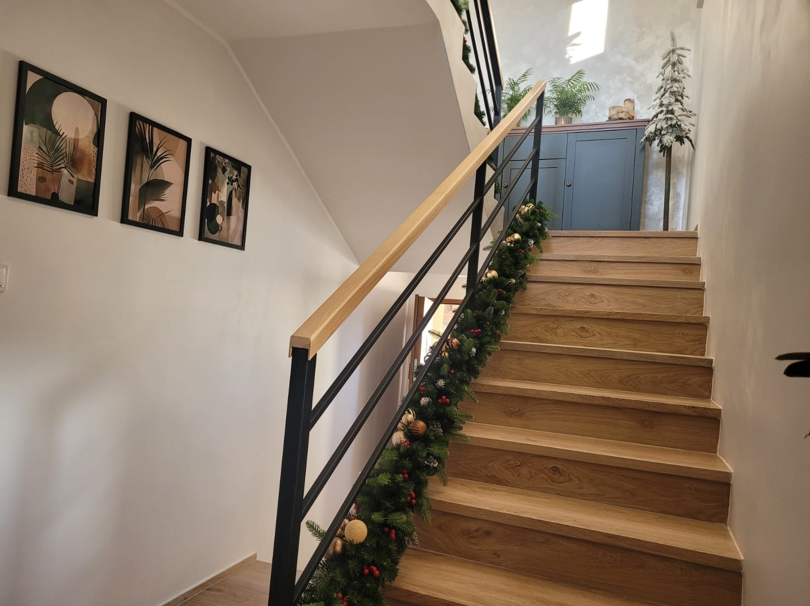 Drewniane schody z czarną metalową balustradą i jasną drewnianą poręczą, ozdobione świąteczną girlandą, widok z dołu na górę, w tle komoda i dekoracje.