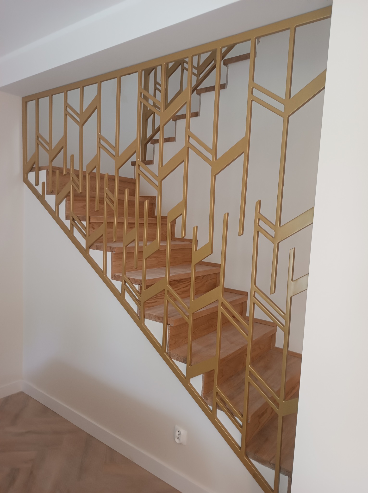 Nowoczesne schody z drewnianymi stopniami i złotą, geometryczną balustradą. Minimalistyczny design, jasne wnętrze, subtelne oświetlenie.