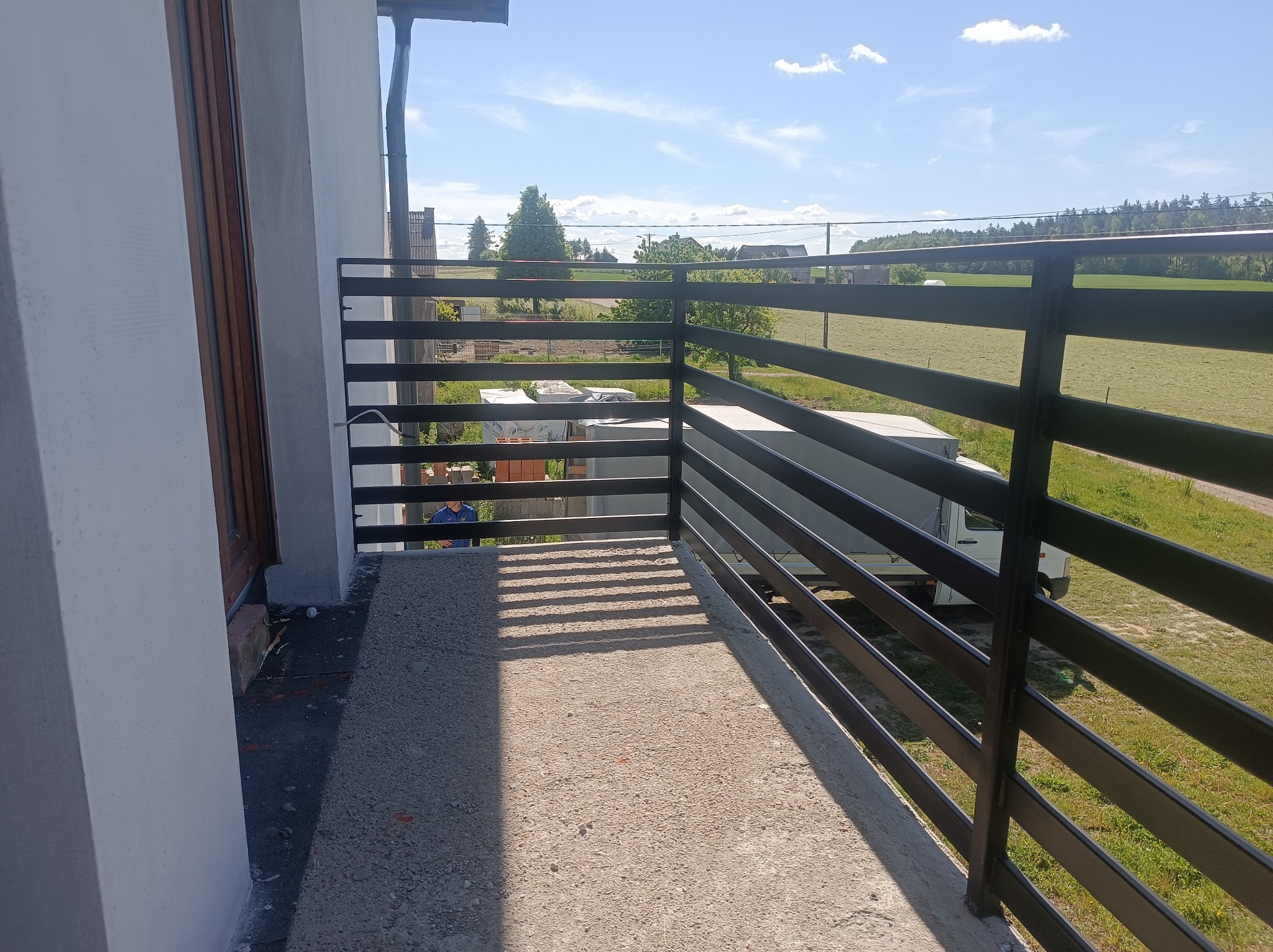 Czarna, pozioma balustrada balkonowa z metalu, z widokiem na zielone pola i błękitne niebo. Detale konstrukcyjne i perspektywa z balkonu.