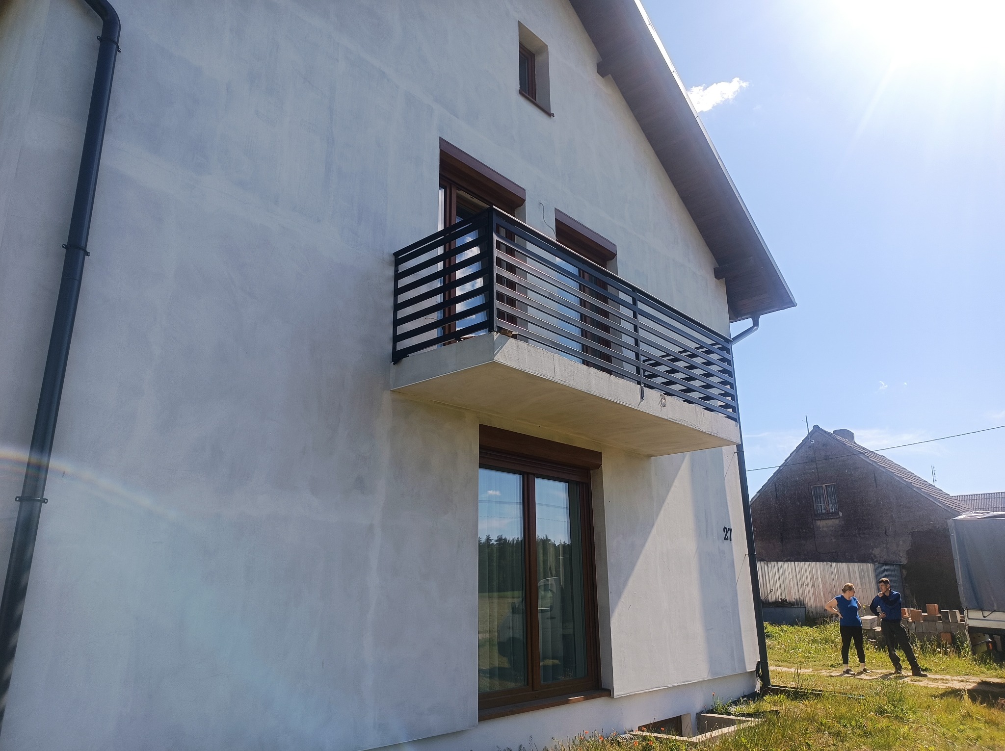 Nowoczesna, czarna balustrada balkonowa z poziomych profili na tle jasnej elewacji domu z brązowymi oknami i roletami; widok z dołu w słoneczny dzień.