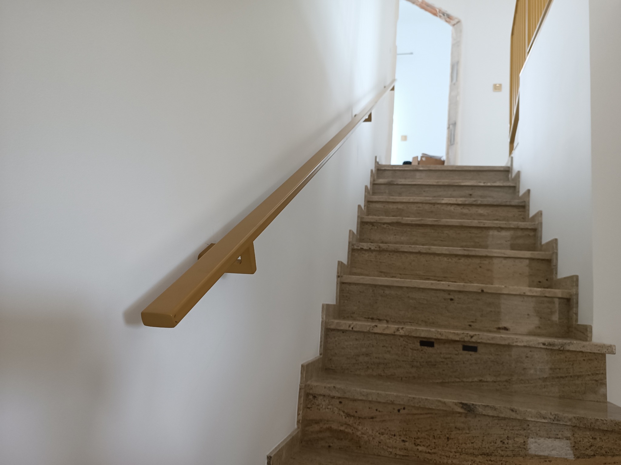 Schody z kamienia z beżową poręczą zamontowaną na ścianie. Widok z dołu, w tle otwór drzwiowy i fragment balustrady.