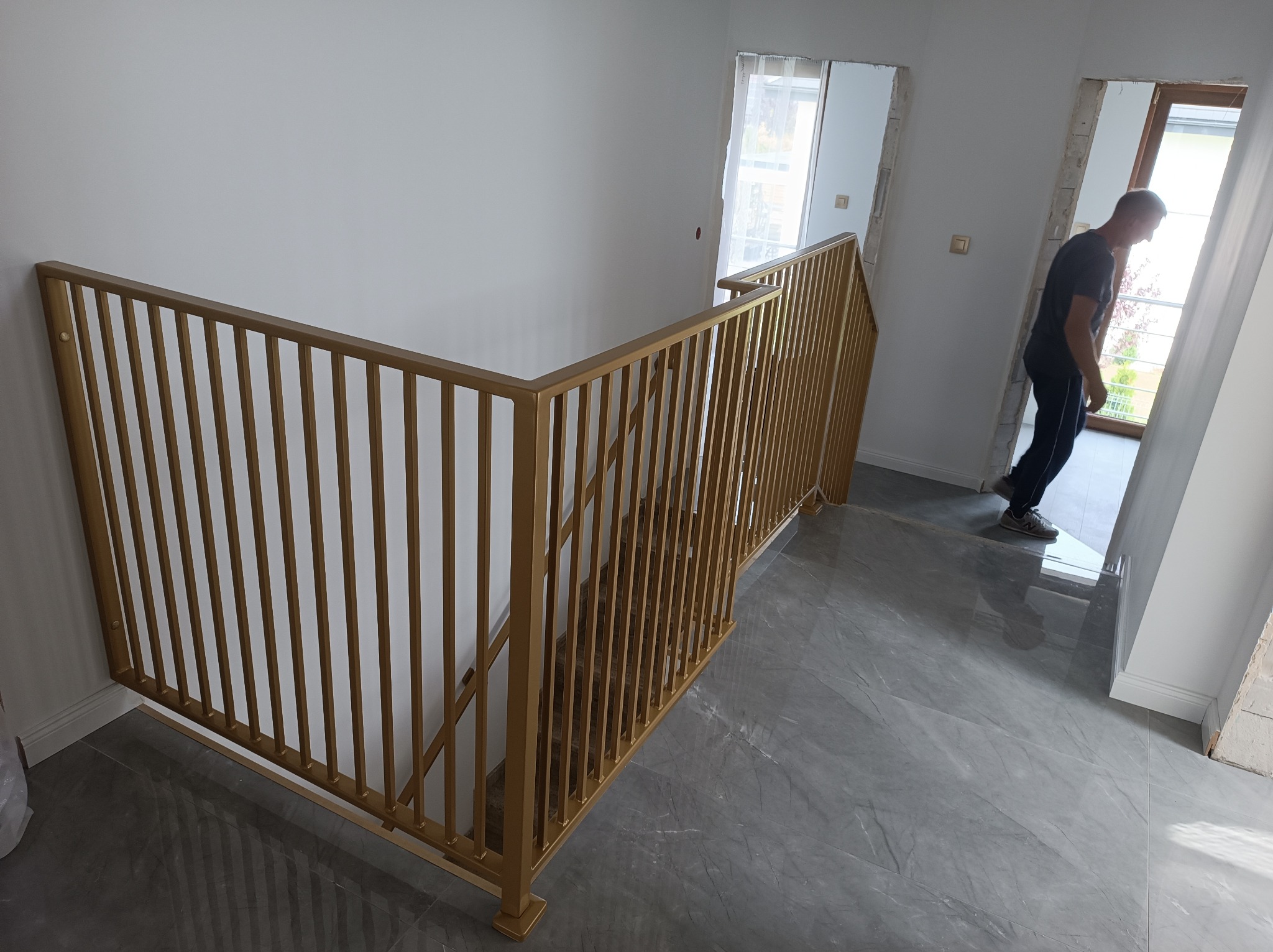 Złota, metalowa konstrukcja schodowa z pionowymi szczebelkami w nowoczesnym wnętrzu. Widoczny fragment schodów i osoba w przejściu.