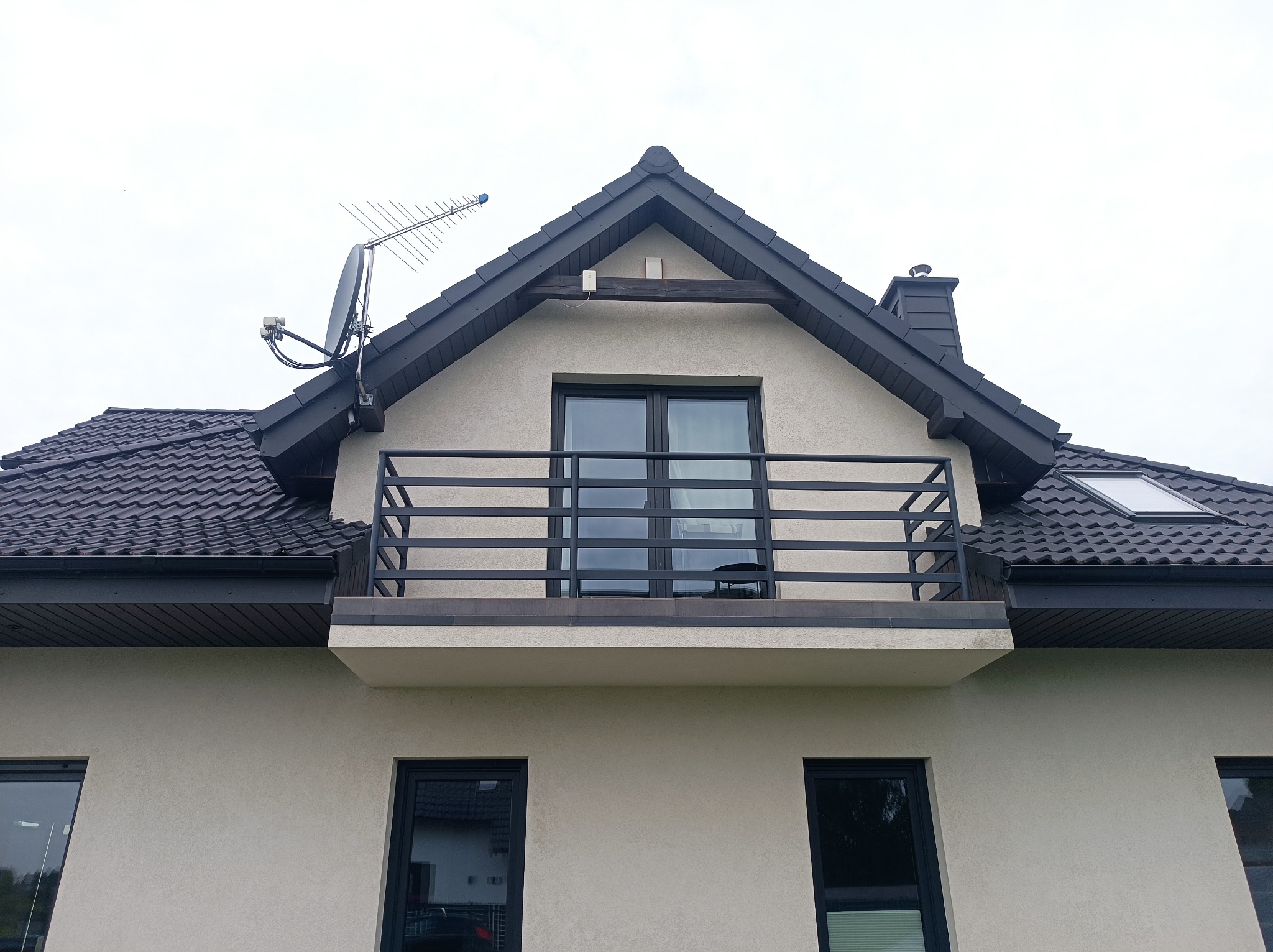 Dom z balkonem zabezpieczonym metalową, czarną balustradą. Na dachu widoczna antena satelitarna i komin. Okna i dach w ciemnej tonacji, elewacja jasna.