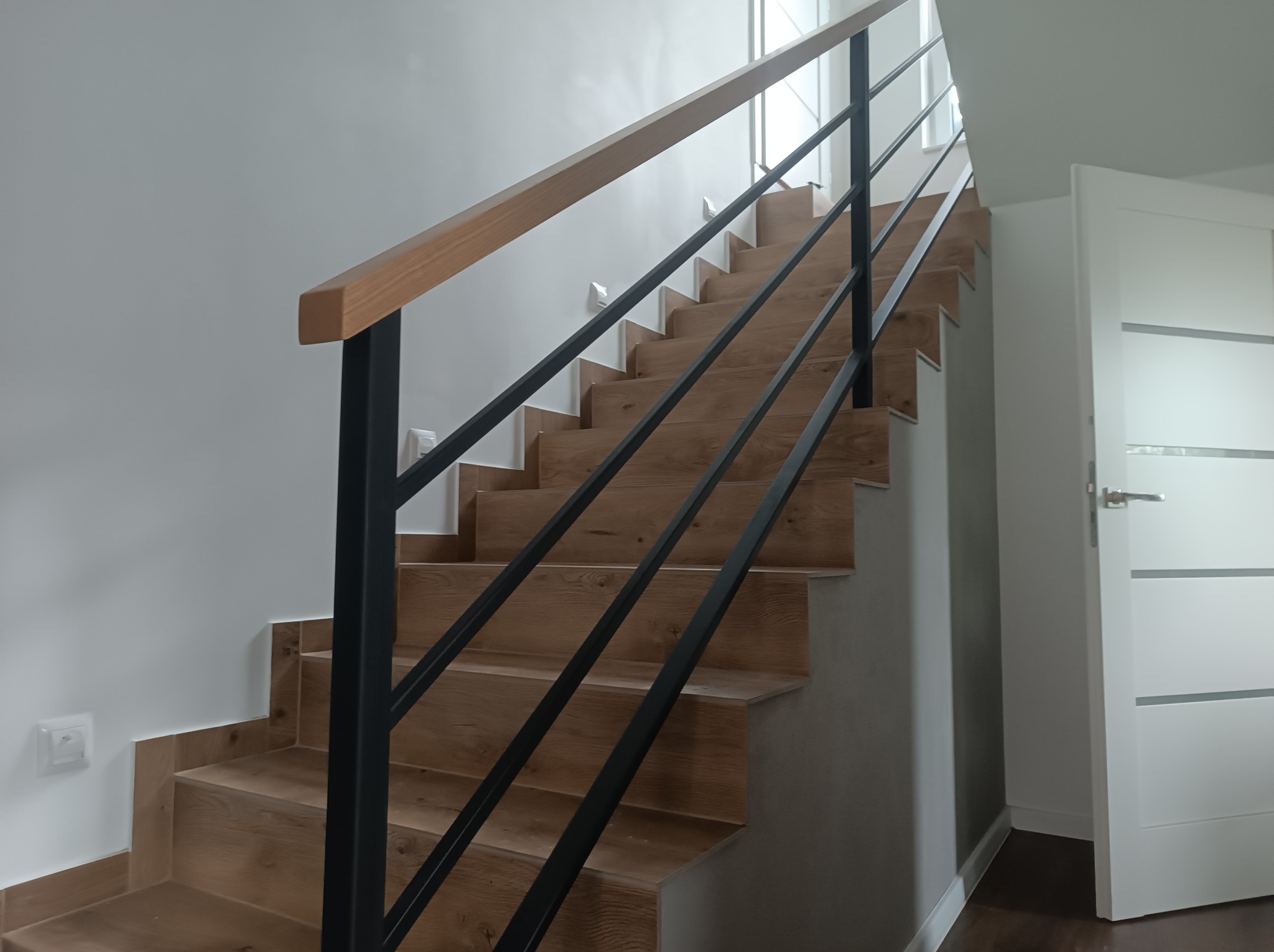 Nowoczesne schody z drewnianymi stopniami i czarną, minimalistyczną balustradą. Jasne wnętrze z białą ścianą i drzwiami. Widok z dołu, akcent na detal wykończenia.