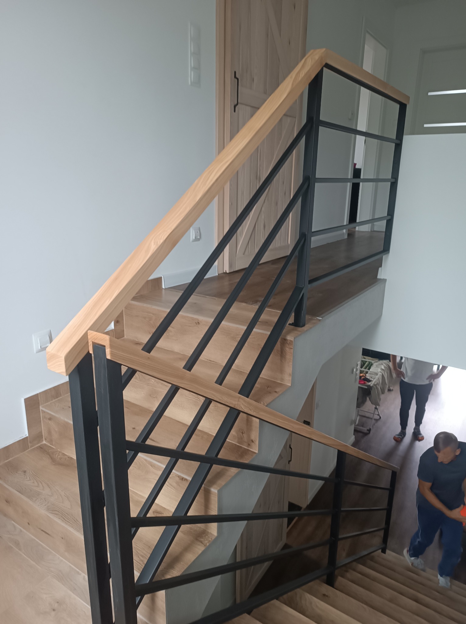 Nowoczesne schody z drewnianymi stopniami i czarną, metalową balustradą z drewnianą poręczą. Kąt ujęcia z góry, widoczne fragmenty osób na schodach.