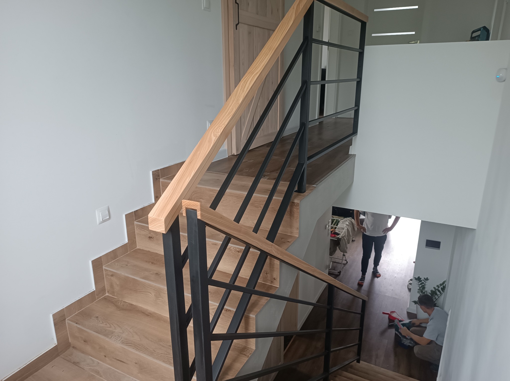 Nowoczesne schody z drewnianymi stopniami i czarną, metalową balustradą, widoczne z góry. W tle osoby na piętrze i roślina doniczkowa.