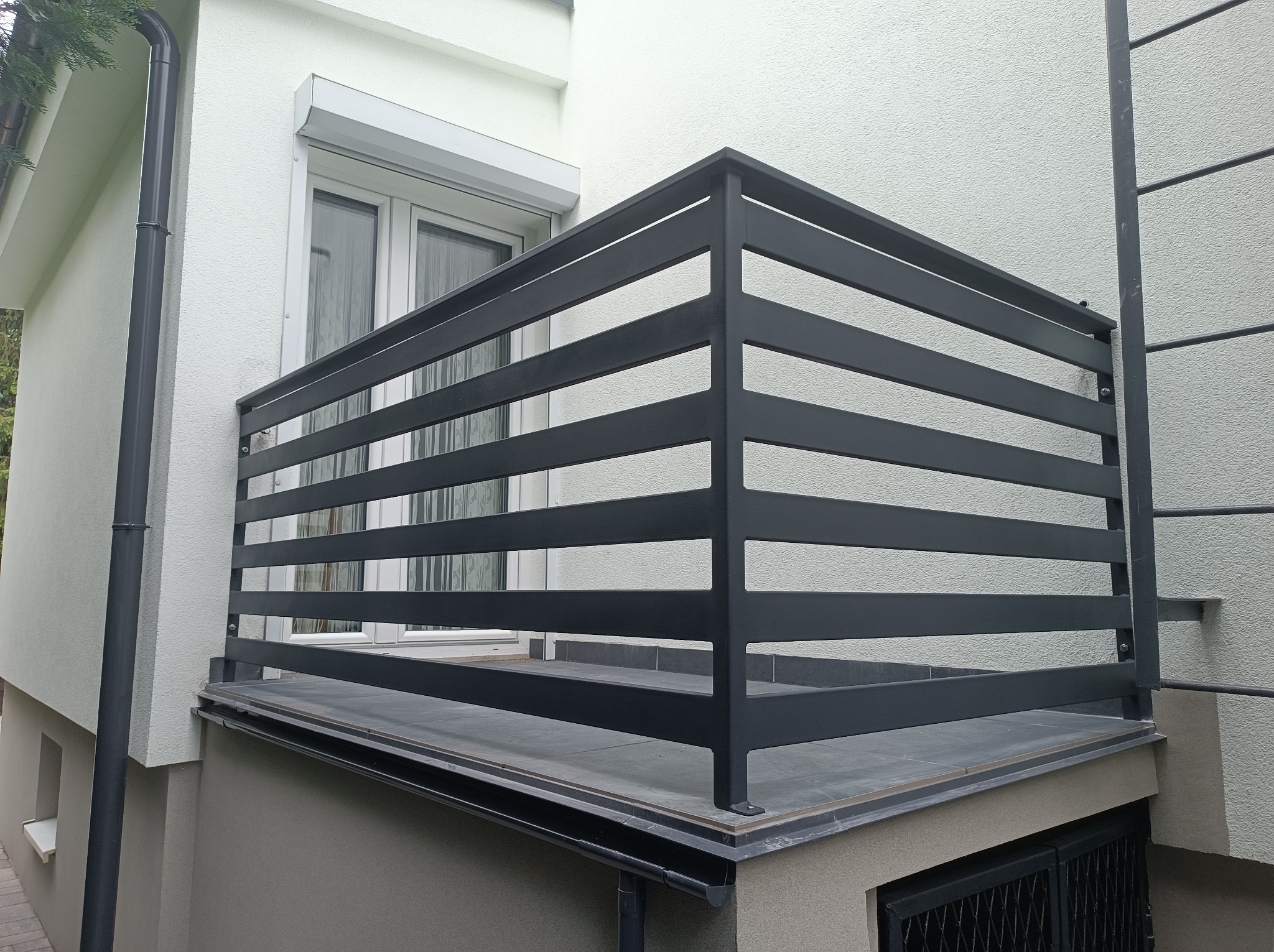 Nowoczesna, szara balustrada balkonowa z poziomych profili, zamontowana na płytkach. Widok z dołu, fragment elewacji budynku z oknem i roletą.