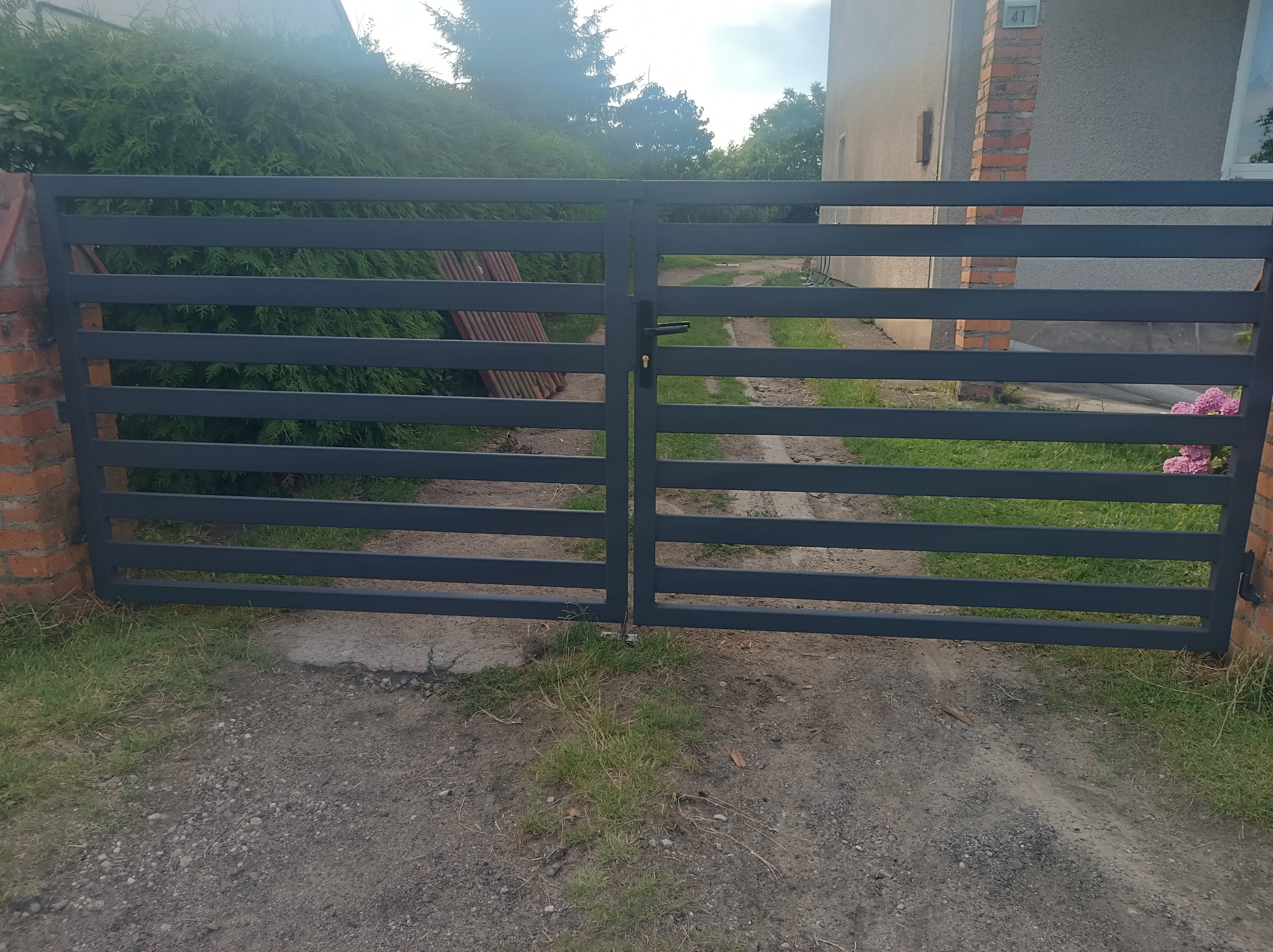 Nowoczesna, pozioma brama wjazdowa w kolorze grafitowym, zamontowana na ceglanych słupkach. Widoczne zarośla w tle, numer domu 41.