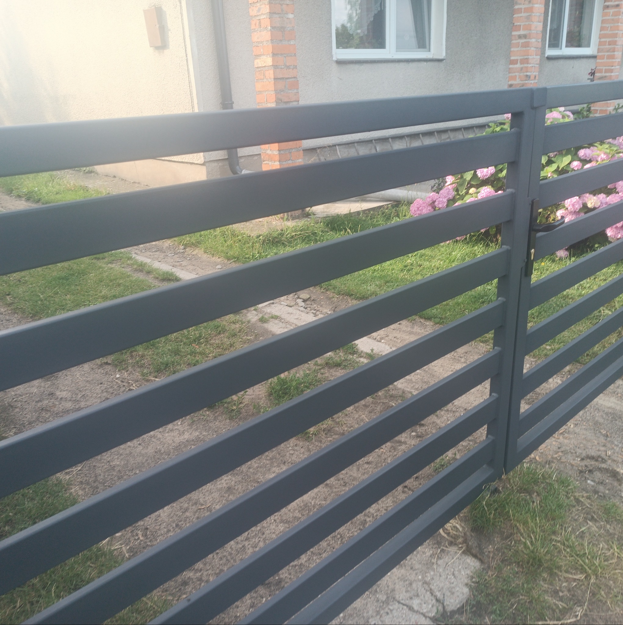 Nowoczesna, pozioma brama wjazdowa w szarym kolorze, z widocznymi kwiatami hortensji w tle, umieszczona przed domem z elementami ceglanymi.