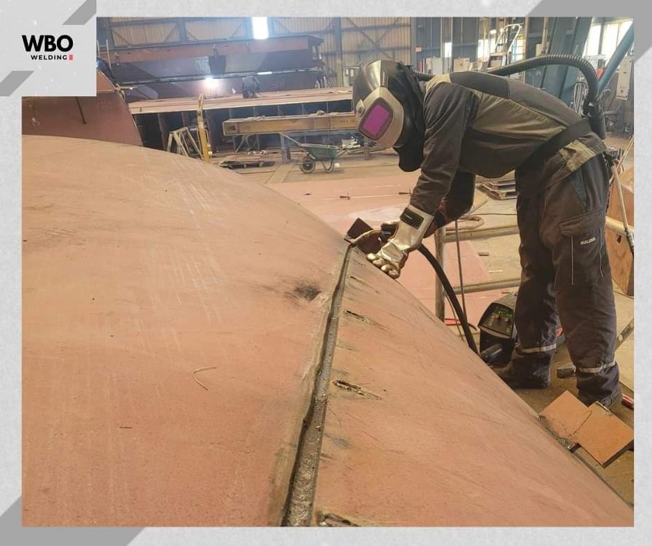 Pracownik w kombinezonie spawalniczym łączy dwie duże, zakrzywione płyty metalowe. Widoczny spaw, sprzęt spawalniczy i fragment hali produkcyjnej.