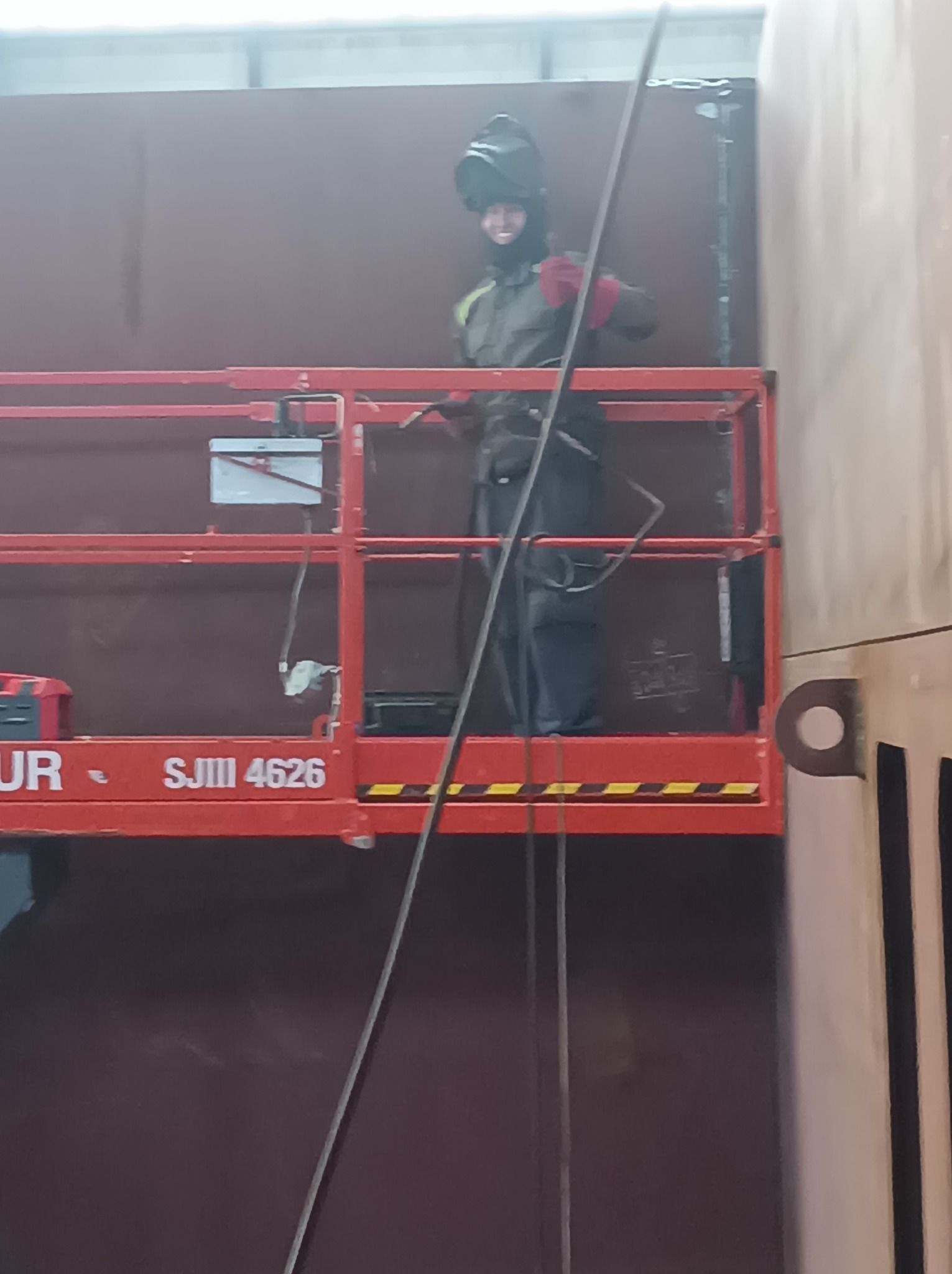 Pracownik w masce spawalniczej na podnośniku, wykonuje prace spawalnicze na dużej, metalowej konstrukcji w hali. Widoczne elementy konstrukcji i narzędzia.