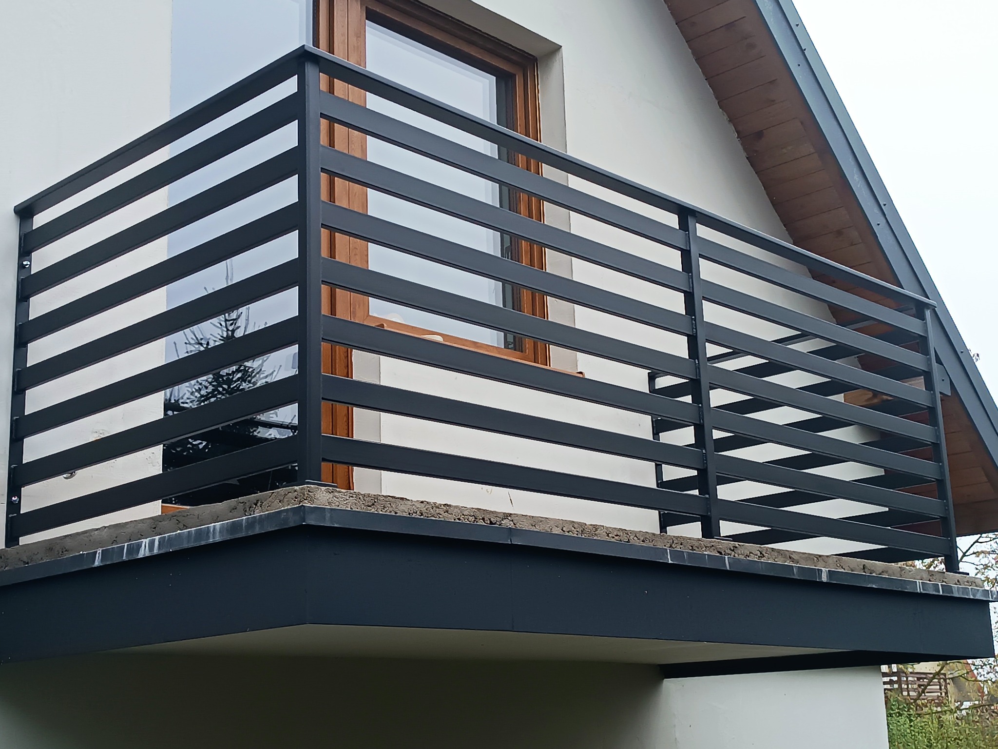 Nowoczesna, pozioma, czarna konstrukcja na balkonie z widocznym oknem i fragmentem dachu. Minimalistyczny design z wyraźnymi liniami i kątami.