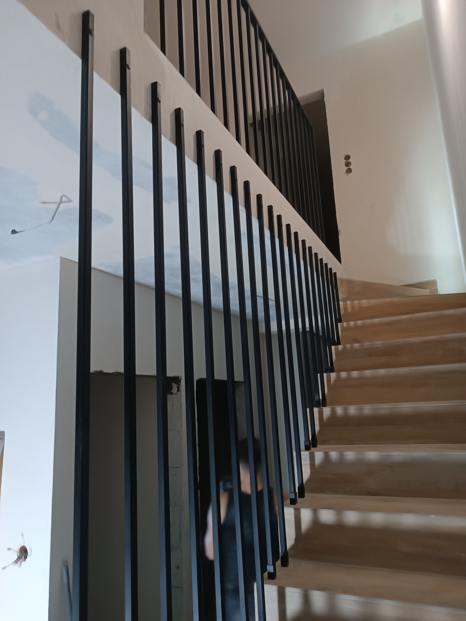 Nowoczesne schody drewniane z czarną, metalową balustradą o minimalistycznym designie. Widok z dołu, podkreślający pionowe linie balustrady i jasne drewno stopni.