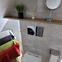 Nowoczesna łazienka na poddaszu z szarą płytką, podwieszaną toaletą, chromowanym przyciskiem spłukującym, półką z dekoracjami i suszarką na ręczniki z kolorowymi ręcznikami.