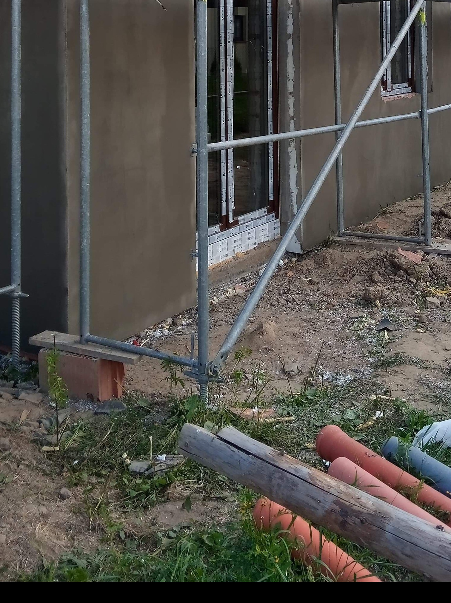 Surowa elewacja budynku w trakcie tynkowania, widoczne rusztowania, okna z folią ochronną i rury PCV na ziemi.
