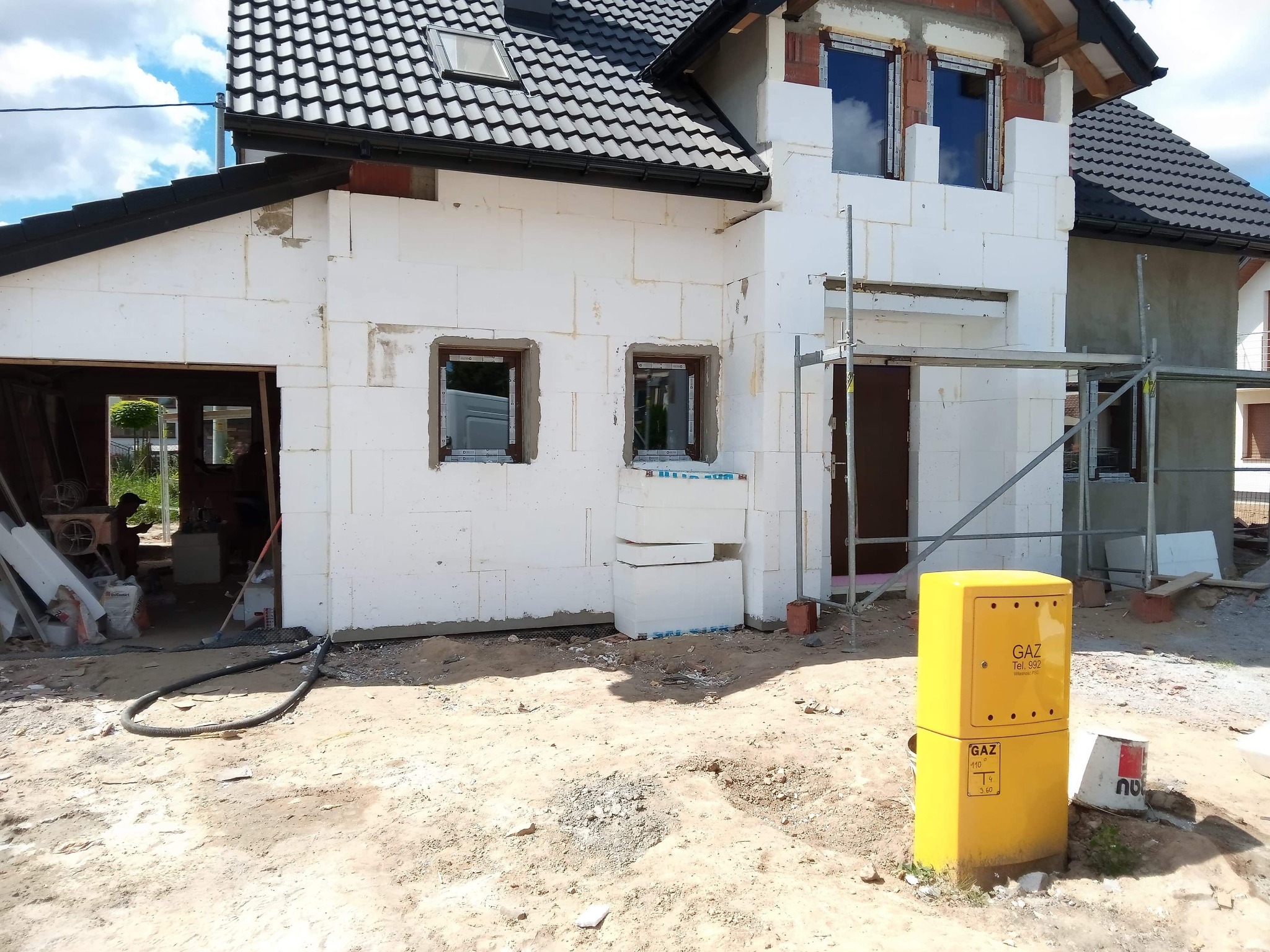 Dom w trakcie ocieplania styropianem, widoczne rusztowania, okna bez parapetów i żółta skrzynka gazowa na pierwszym planie.
