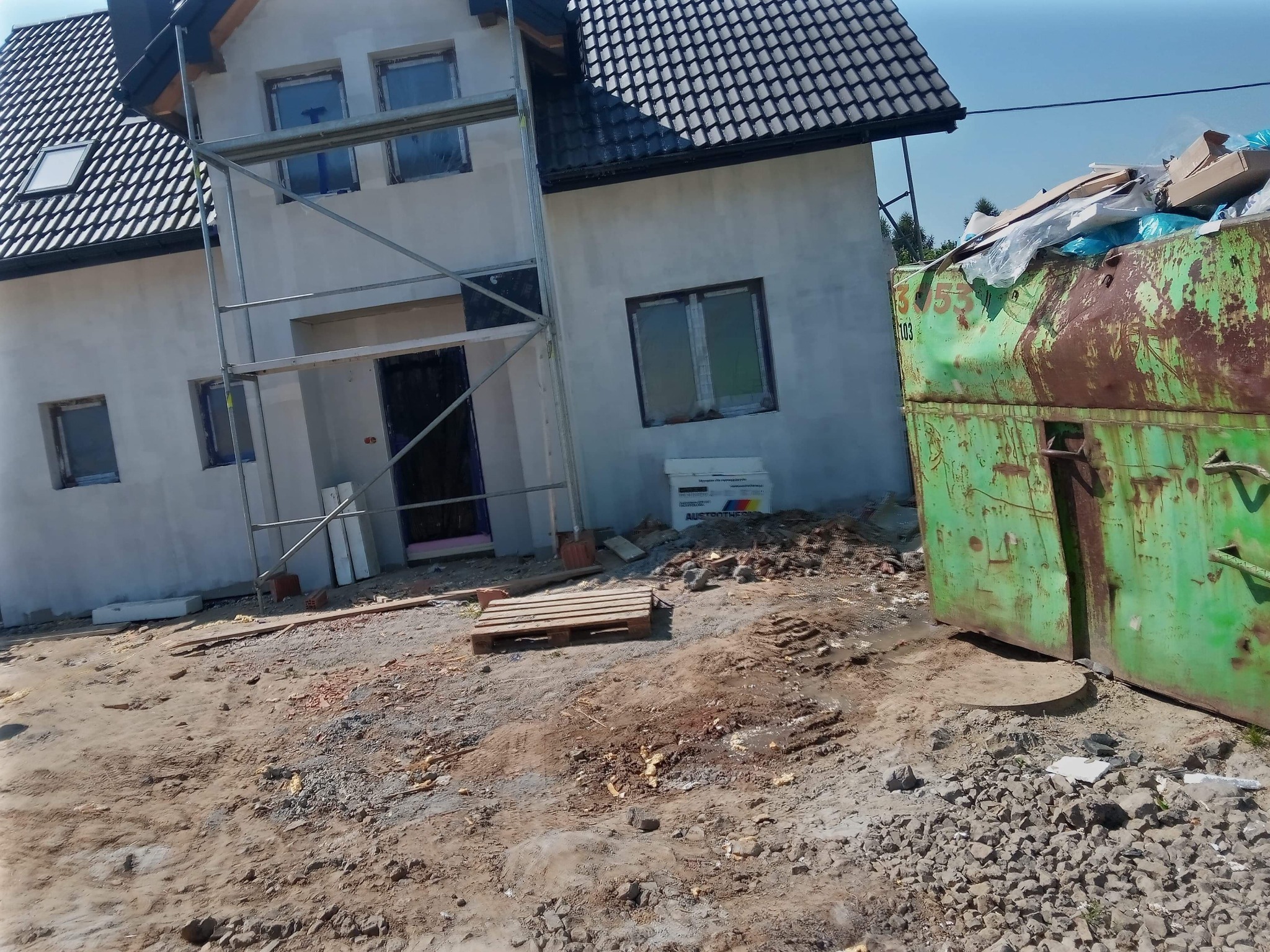 Surowa elewacja domu jednorodzinnego w trakcie prac wykończeniowych, widoczne rusztowanie, paleta i kontener na odpady budowlane.