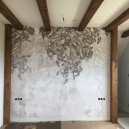 Ściana z dekoracyjną tapetą w odcieniach szarości z motywem roślinnym, drewniane belki stropowe i słupy, przygotowane otwory na gniazdka elektryczne, podłoga częściowo zabezpieczona kartonem.