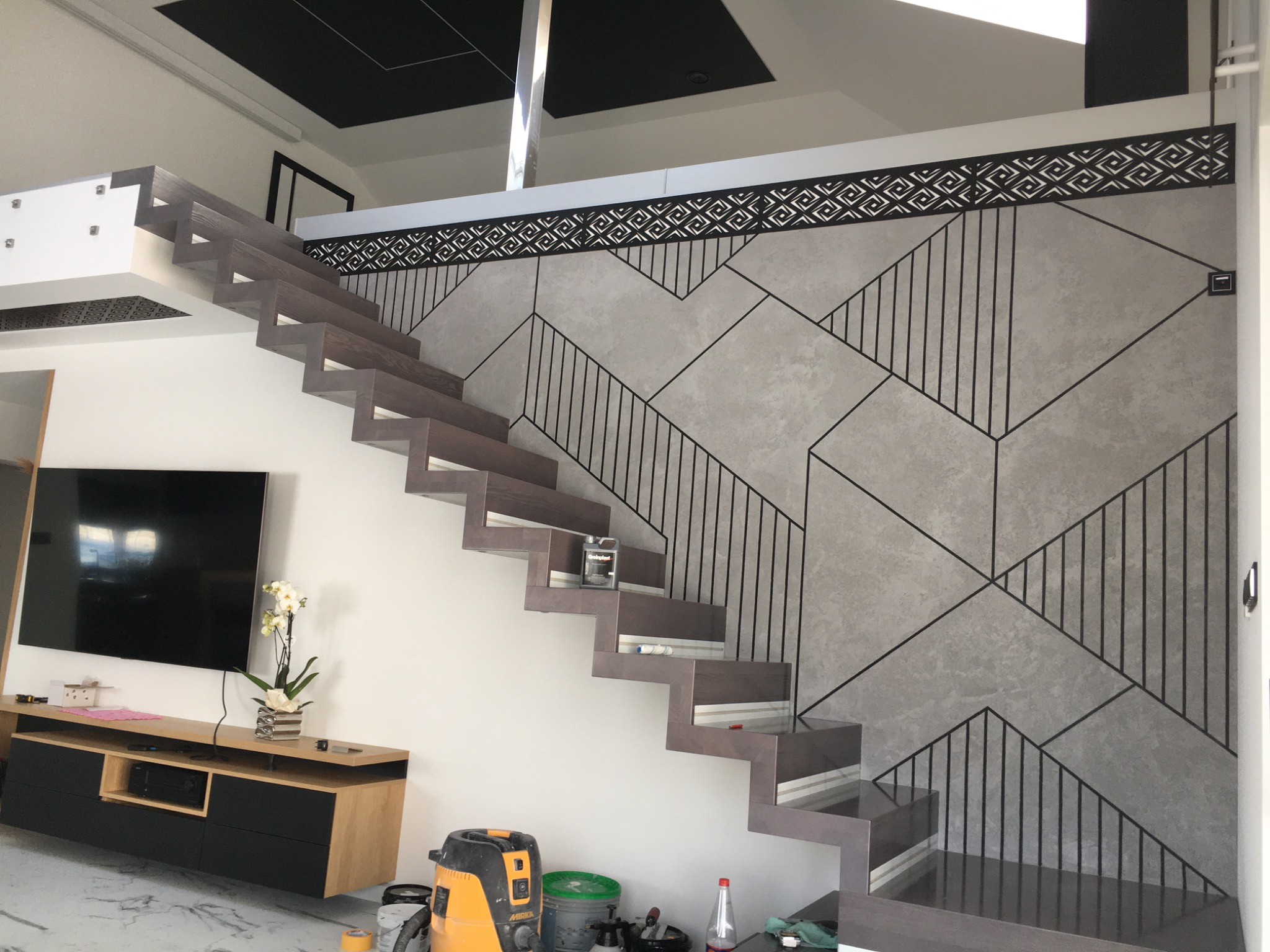 Nowoczesne, ażurowe schody drewniane z podświetleniem LED, prowadzące na antresolę z dekoracyjną ścianą w geometryczne wzory, widoczny odkurzacz i akcesoria malarskie na podłodze.