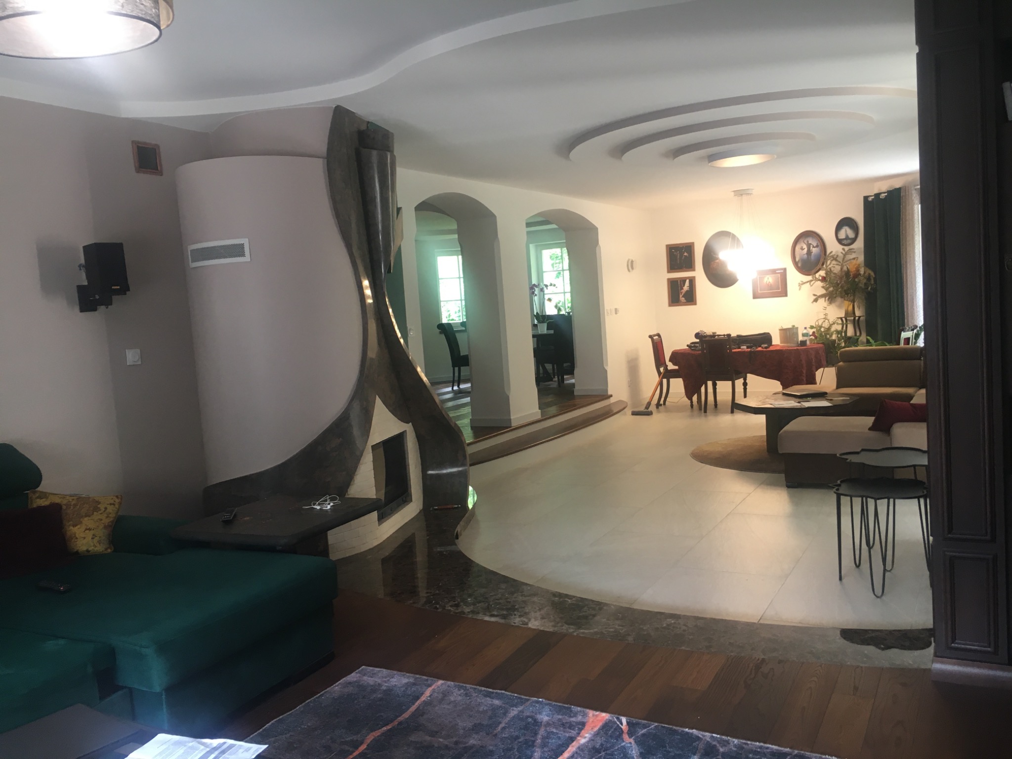 Przestronny salon z nowoczesnym kominkiem o nietypowym kształcie, oddzielającym strefę wypoczynkową od jadalni, widoczny fragment zielonej sofy i drewniana podłoga przechodząca w jasne płytki.