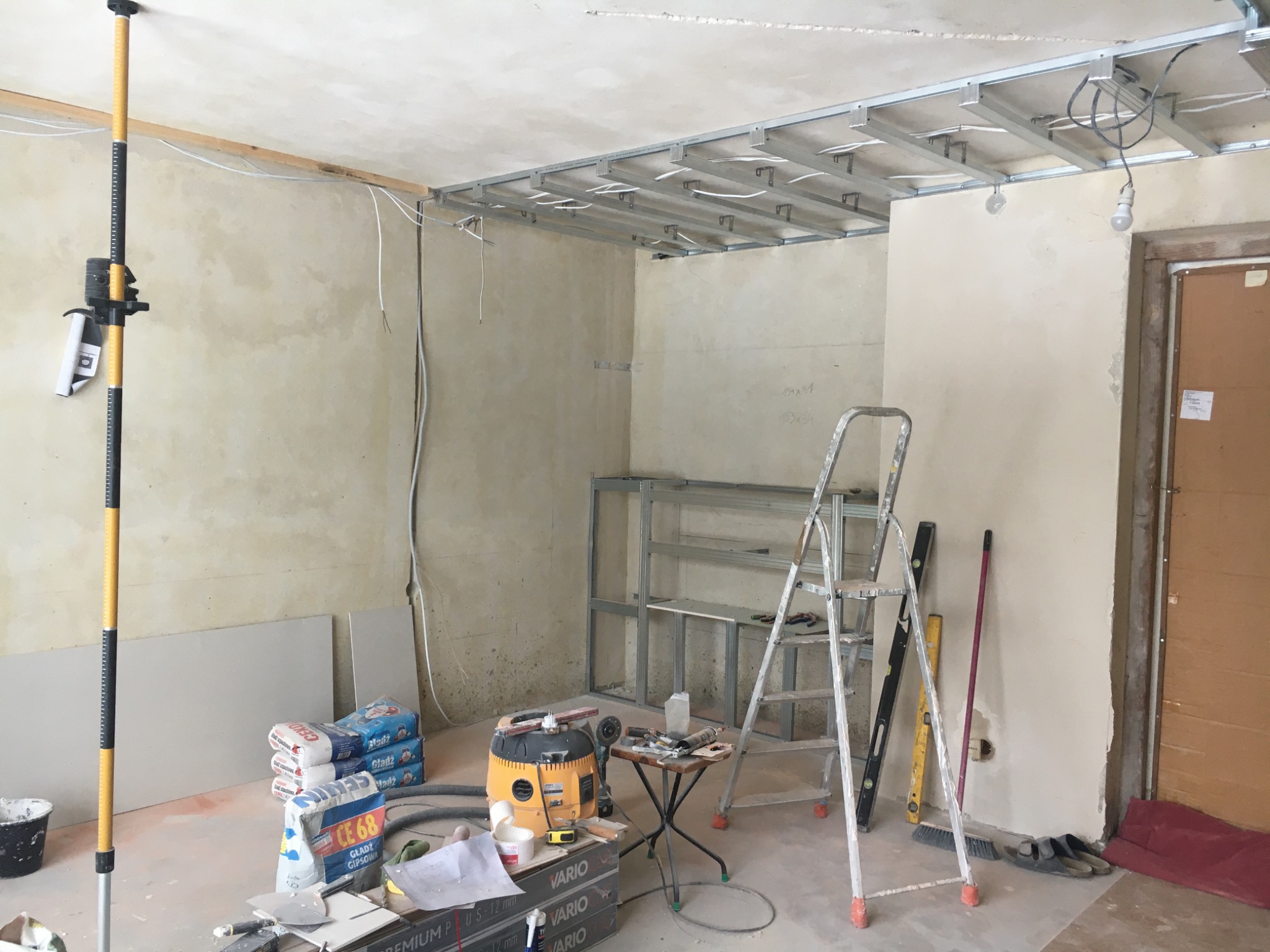 Montaż metalowej konstrukcji sufitu podwieszanego w trakcie remontu, widoczne profile, przewody elektryczne, laserowy niwelator, worki z gładzią gipsową, drabina i narzędzia.
