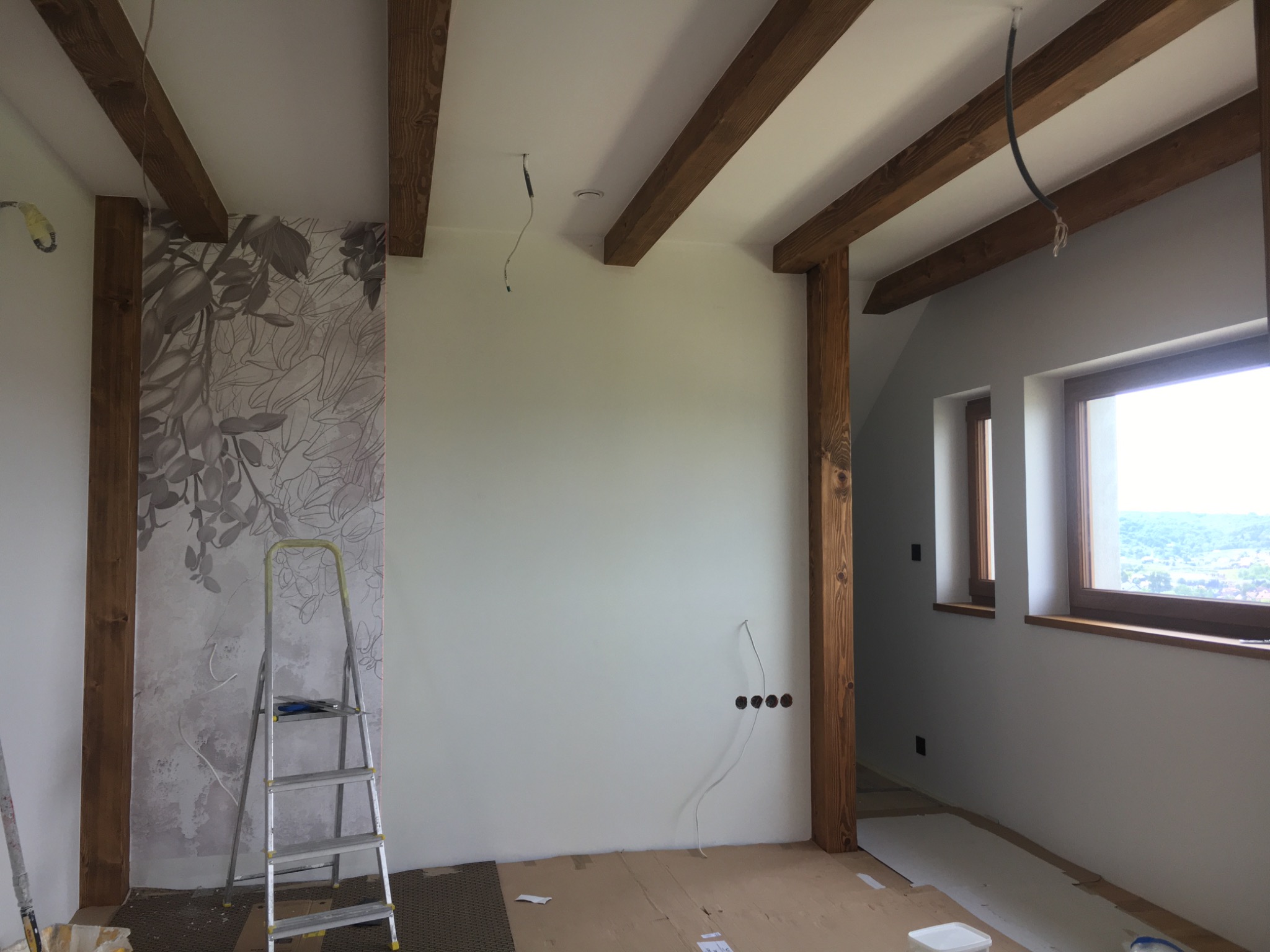 Pokój w trakcie wykańczania z drewnianymi belkami stropowymi, częściowo wytapetowaną ścianą z motywem roślinnym i rozłożonymi kartonami na podłodze, widoczne gniazdka elektryczne i drabina aluminiowa.