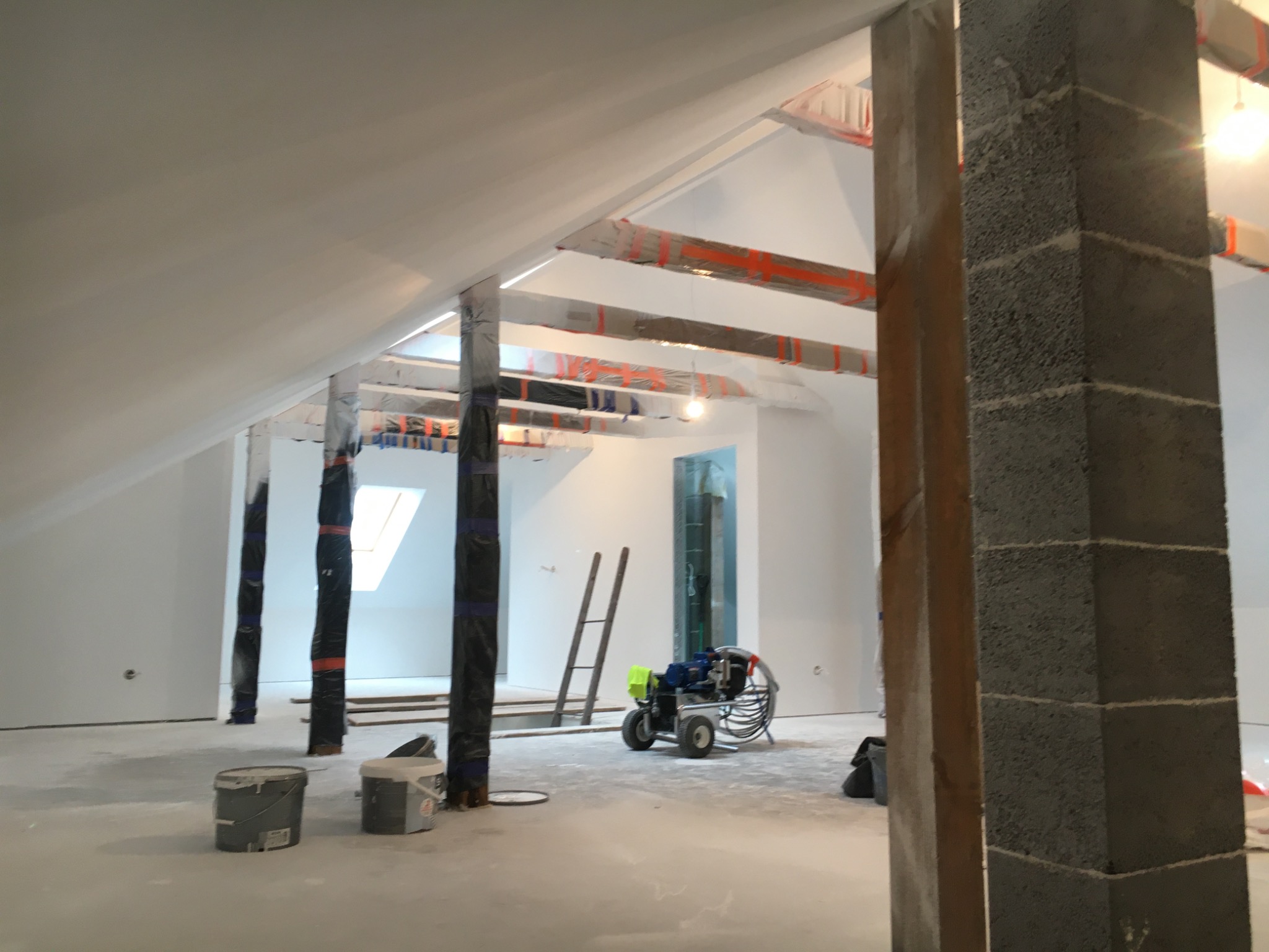 Poddasze w trakcie prac wykończeniowych: widoczne otynkowane ściany, zabezpieczone folią słupy, drabina oparta o ścianę oraz agregat malarski na podłodze pokrytej świeżym tynkiem.
