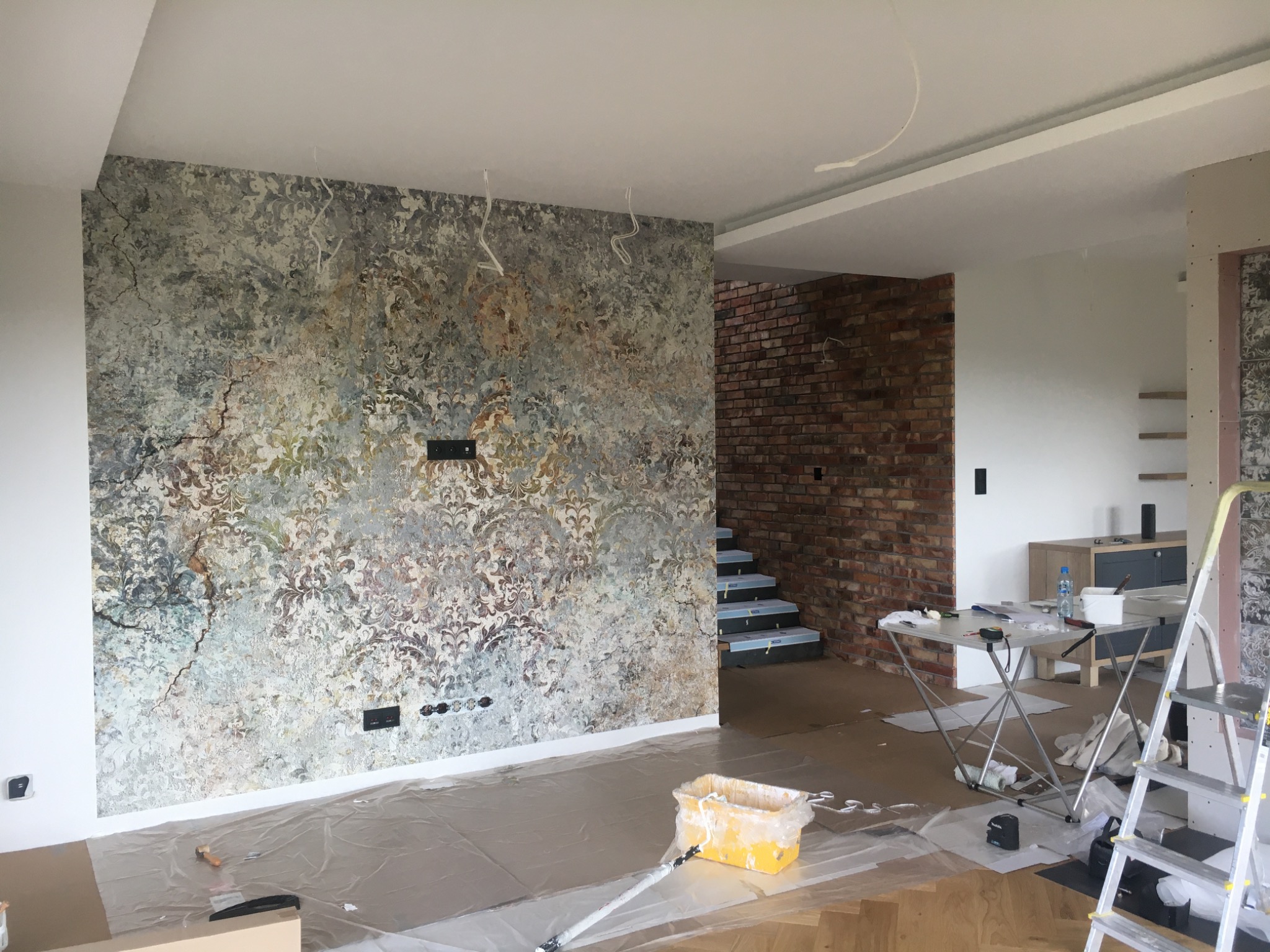 Wnętrze w trakcie prac: ściana z wzorzystą tapetą, zabezpieczona folią podłoga, żółte wiadro z farbą i wałkiem, widoczne gniazdka elektryczne, w tle schody z ceglaną ścianą.