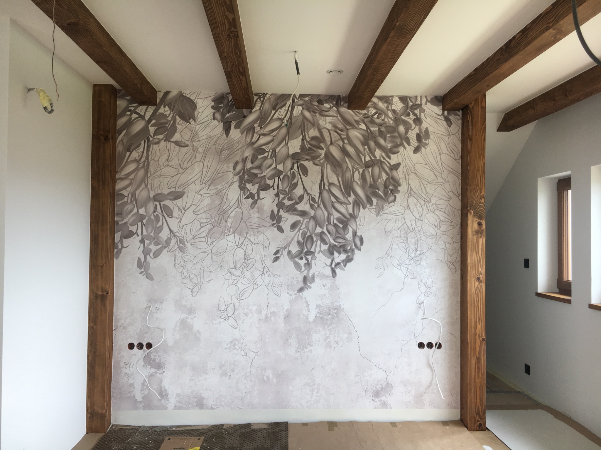Ściana z dekoracyjną tapetą w odcieniach szarości z motywem roślinnym, drewniane belki stropowe i słupy, przygotowane otwory na gniazdka elektryczne, podłoga częściowo zabezpieczona kartonem.
