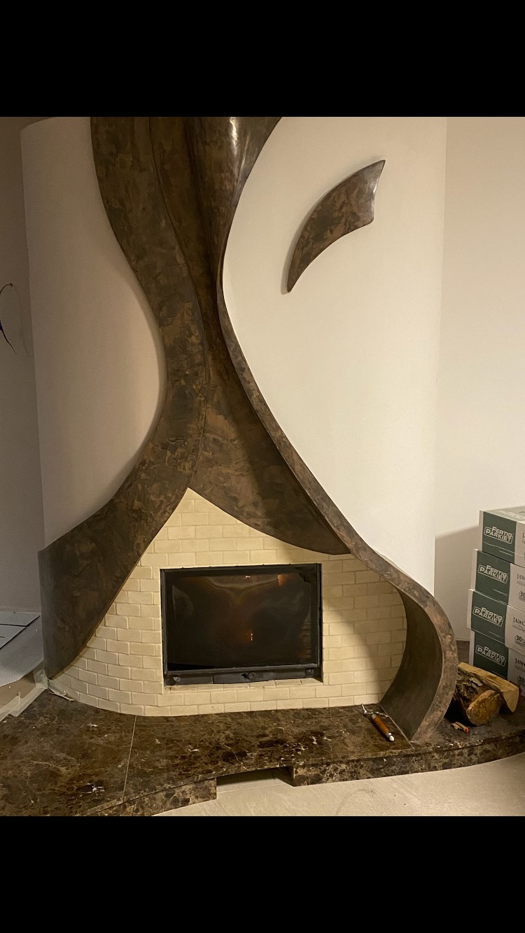 Nowoczesny kominek narożny z obudową w odcieniach brązu i beżu, wykończony marmurem, z widocznymi kartonami z parkietem w tle.