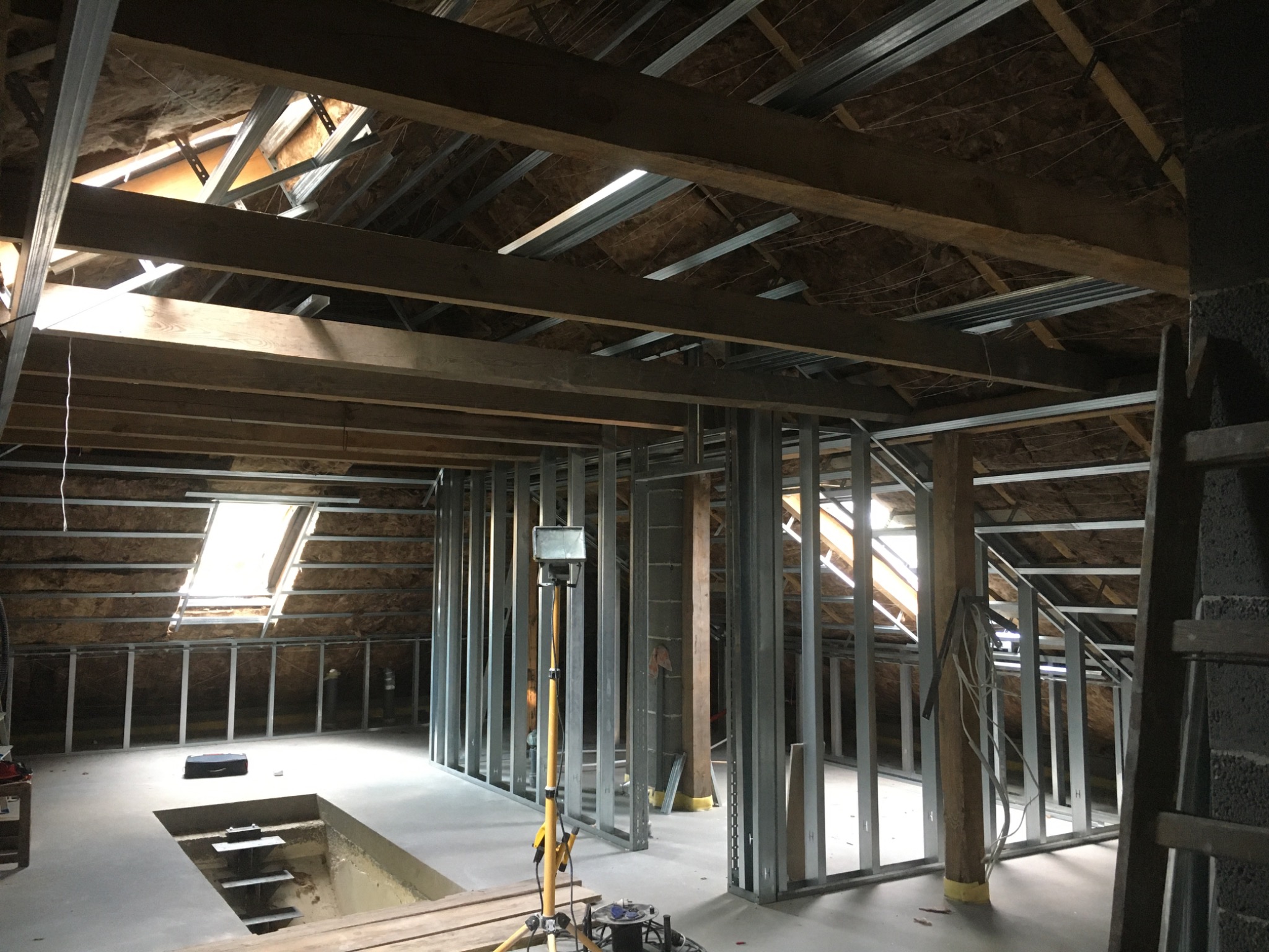 Poddasze w trakcie adaptacji, widoczne metalowe profile konstrukcyjne ścian działowych, drewniana konstrukcja dachu z oknami połaciowymi i częściowo ocieplona wełną mineralną, oświetlenie budowlane...
