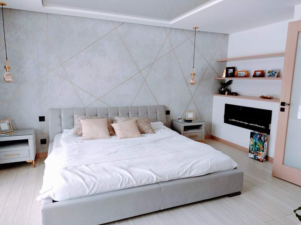 Jasna sypialnia z łóżkiem kontynentalnym, ścianą z szarą fakturą zdobioną złotymi liniami, kominkiem elektrycznym i drewnianą podłogą.