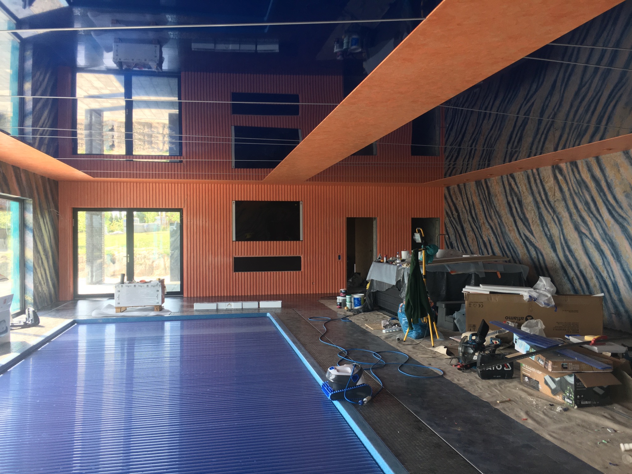 Wnętrze w trakcie remontu z krytym basenem, widoczne pomalowane na pomarańczowo ściany z pionowymi panelami, sufit z połyskiem, częściowo zasłonięty basen z niebieską roletą, oraz narzędzia...