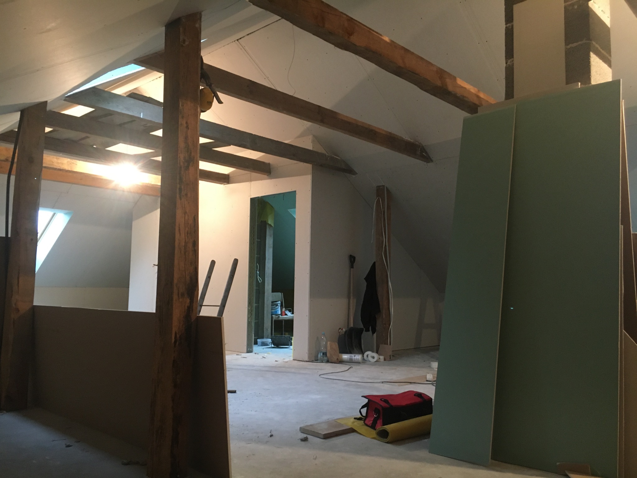 Poddasze w trakcie adaptacji, widoczne drewniane belki konstrukcyjne sufitu, płyty karton-gips, narzędzia i torba narzędziowa na podłodze, etap prac wykończeniowych.