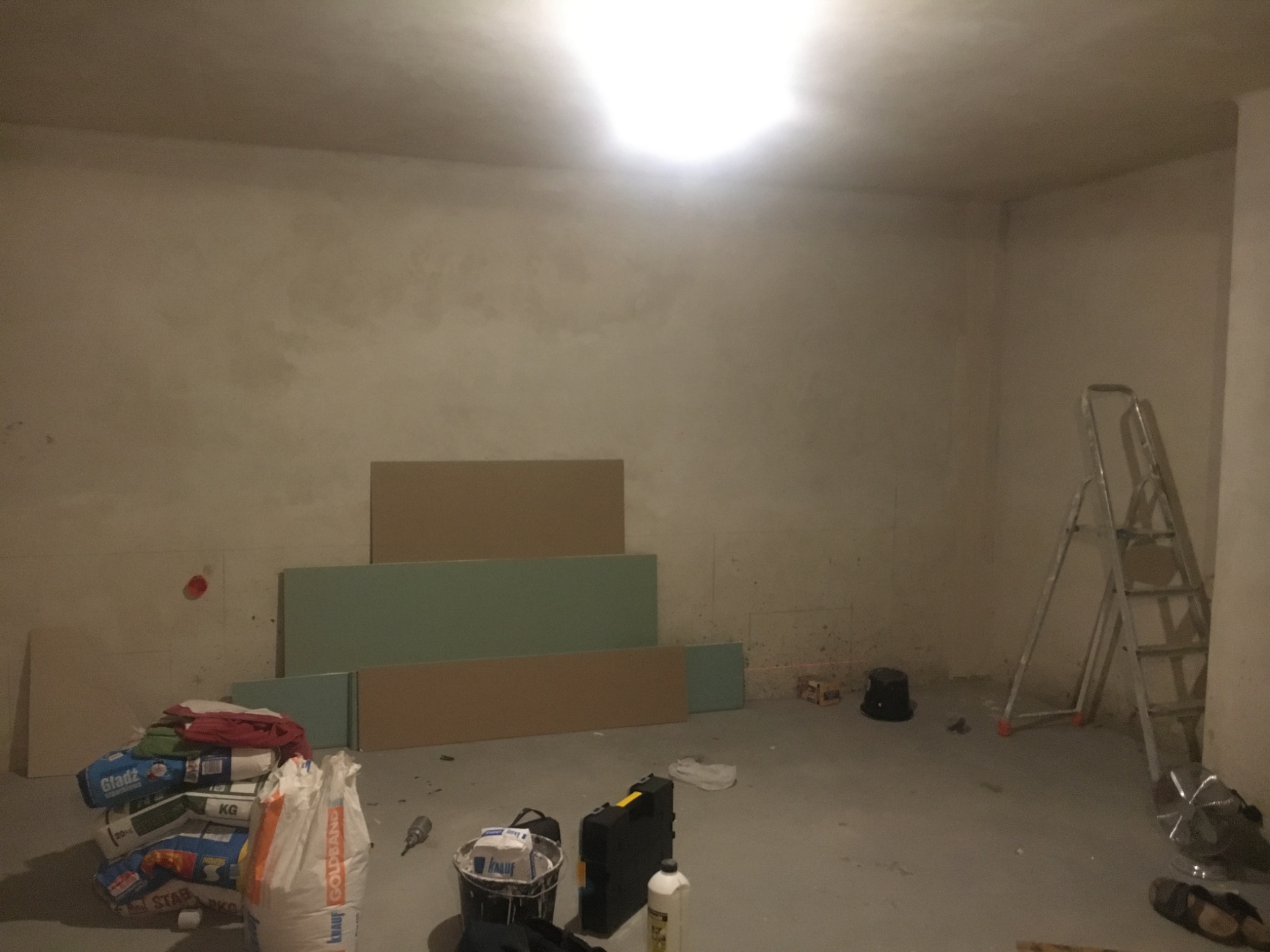 Wnętrze w trakcie prac przygotowawczych: surowe ściany, płyty gipsowo-kartonowe o różnych odcieniach, drabina aluminiowa, worki z zaprawą i narzędzia budowlane rozłożone na podłodze, widoczne ślady...