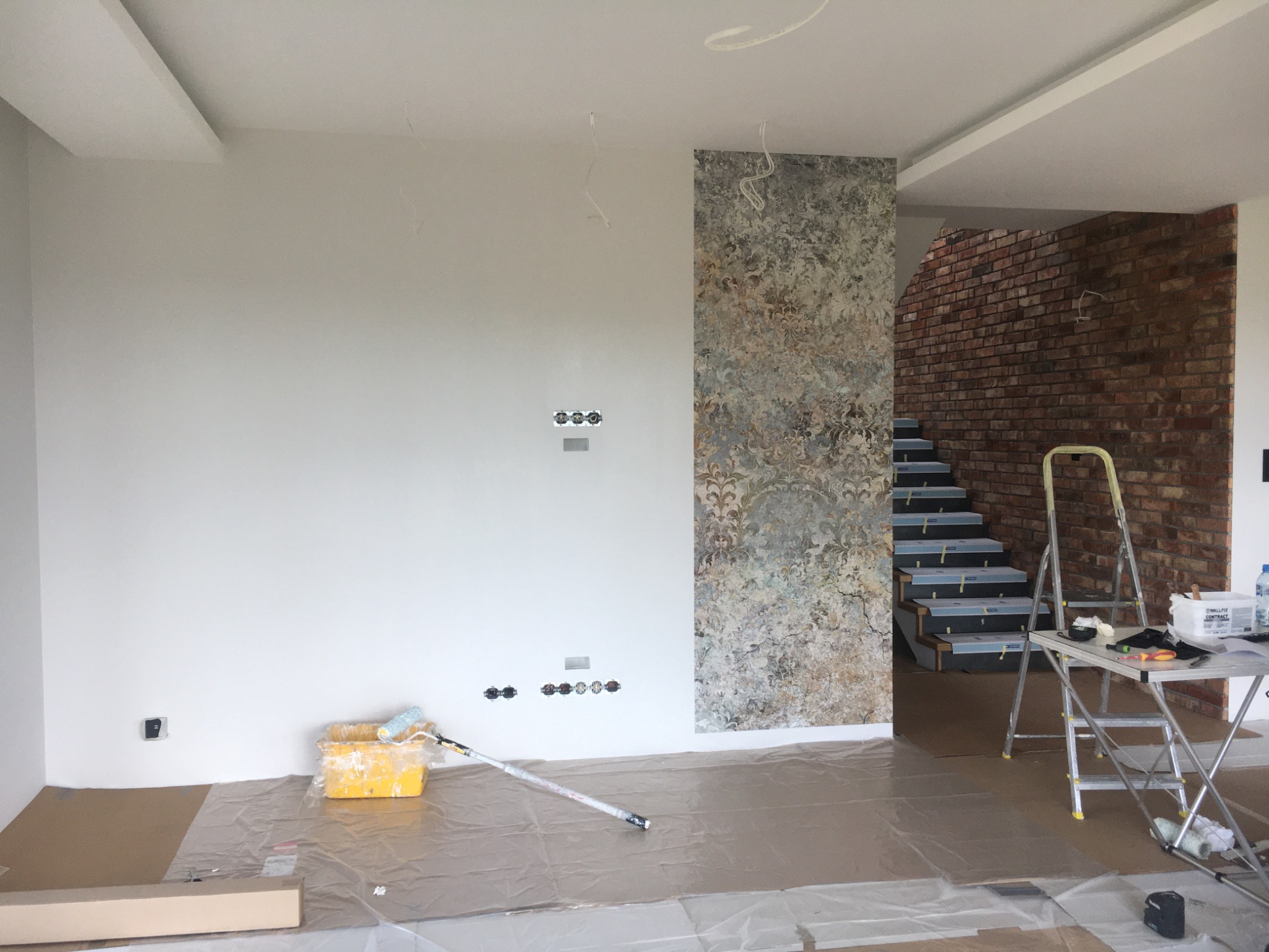 Przygotowania do malowania: zabezpieczona folią podłoga, wałek malarski w kuwecie, drabina rozstawiona w pomieszczeniu z fragmentem ściany wykończonej dekoracyjną tapetą i schodami z ceglaną ścianą...