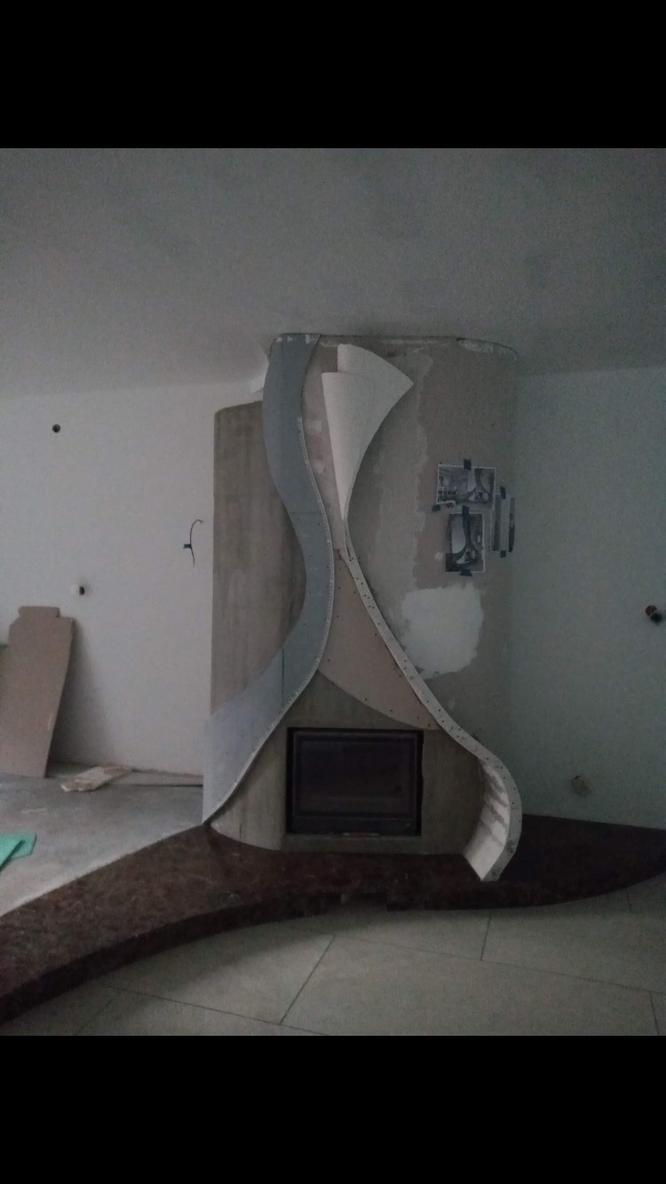 Niedokończona obudowa kominka z płyt gipsowo-kartonowych, o falistym kształcie, z widocznymi śladami szpachlowania i elementami mocującymi, na tle surowych ścian w trakcie prac wykończeniowych.