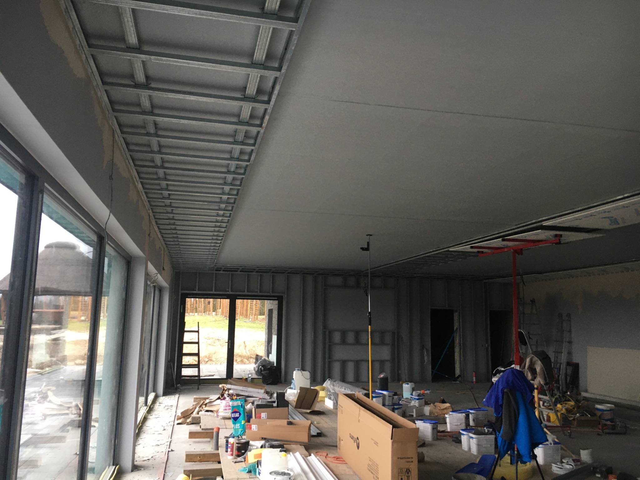 Wnętrze w trakcie prac wykończeniowych: widoczny metalowy stelaż sufitu podwieszanego, częściowo pokryty płytami gipsowo-kartonowymi, liczne puszki z farbą i narzędzia budowlane na podłodze, duże...