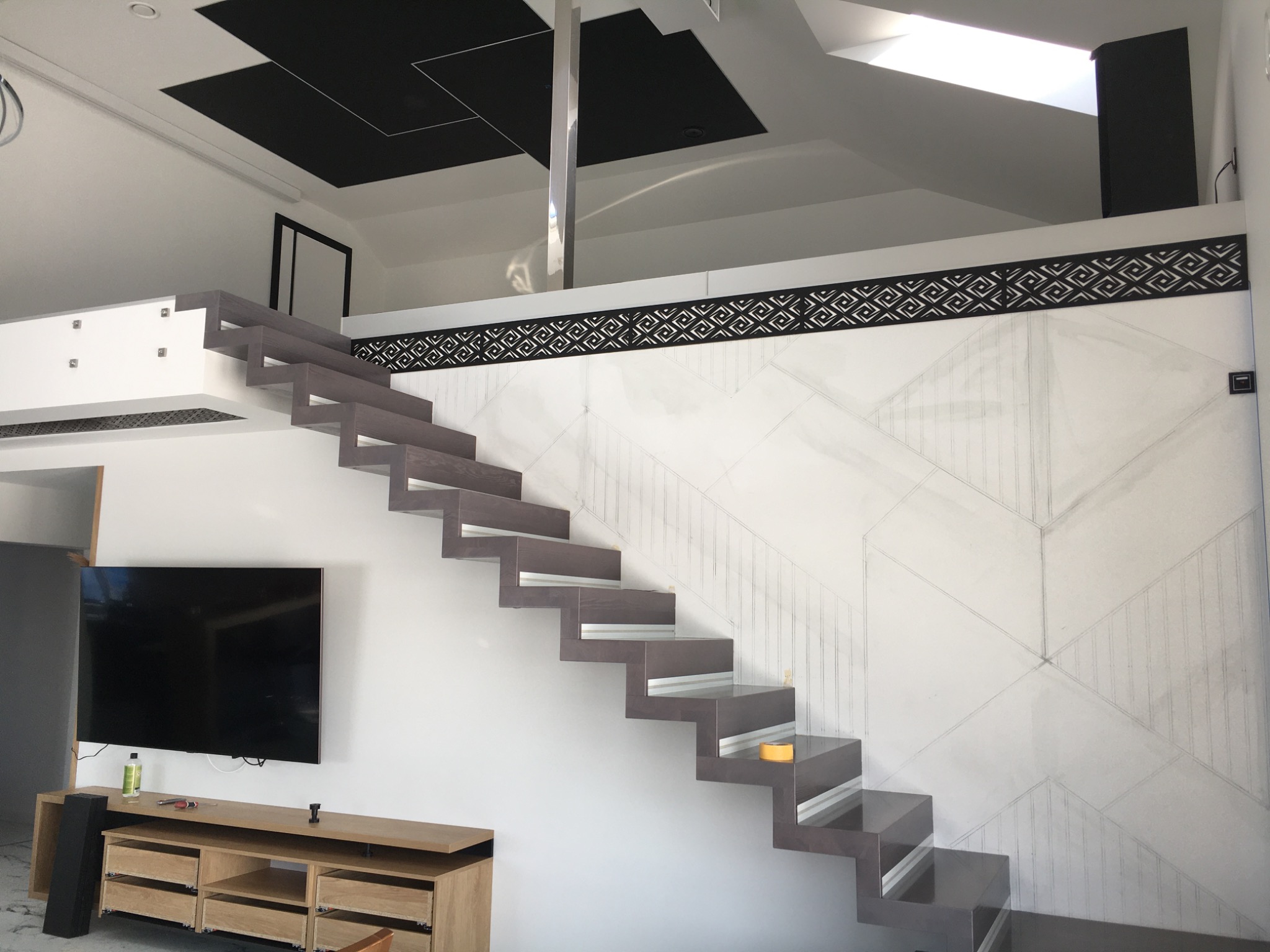 Nowoczesne, wiszące schody drewniane z metalowymi akcentami w jasnym wnętrzu z telewizorem i szafką RTV w trakcie montażu. Ściana z geometrycznym wzorem i dekoracyjnym pasem pod sufitem.