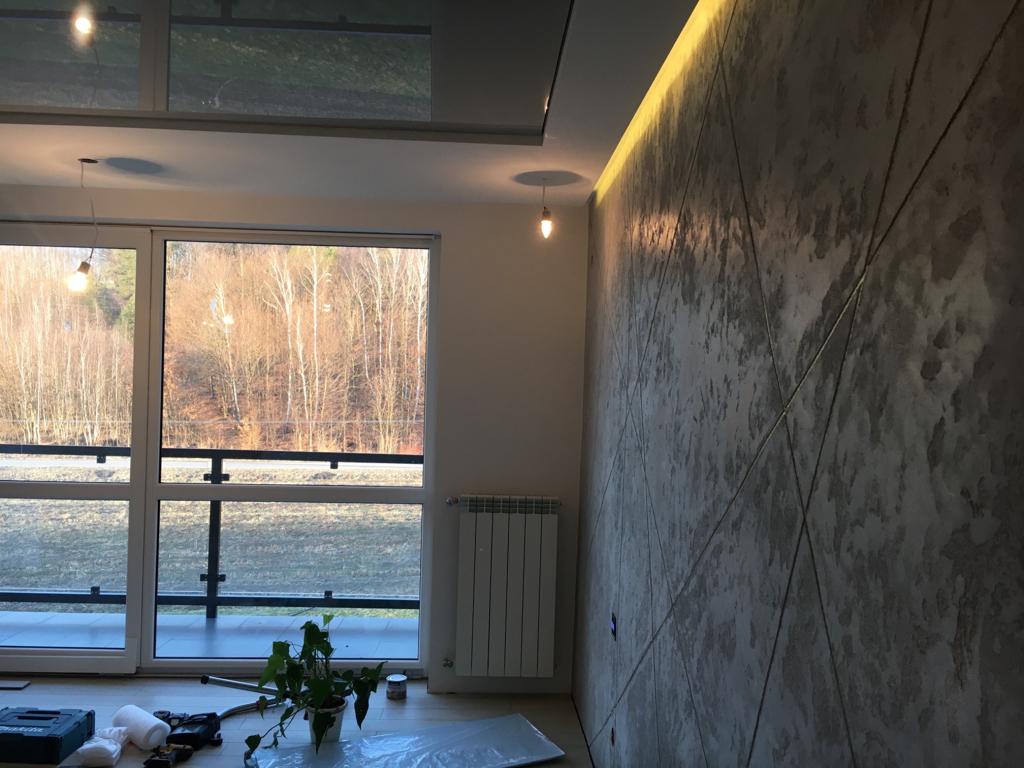 Ściana z dekoracyjną strukturą imitującą beton, podświetlona listwą LED, widok na okno balkonowe z narzędziami i rośliną doniczkową na podłodze.