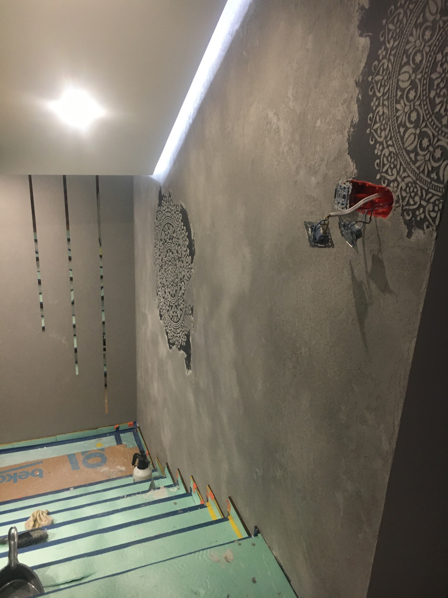 Ściana z szarą gładzią dekoracyjną i wzorem mandali, widoczne gniazdka elektryczne przed montażem, podłoga zabezpieczona folią malarską.