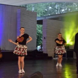 Występ zespołu ludowego na scenie w Łaziskach Górnych, z tancerkami w strojach regionalnych i wokalistą z mikrofonem, oświetlonych reflektorami.