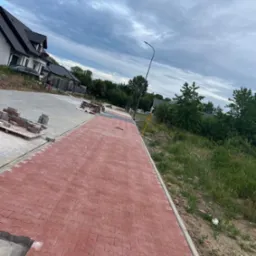 Czerwona kostka brukowa ułożona na długim chodniku wzdłuż ulicy w dzielnicy domów jednorodzinnych, widoczne palety z kostką i trawnik po prawej stronie.