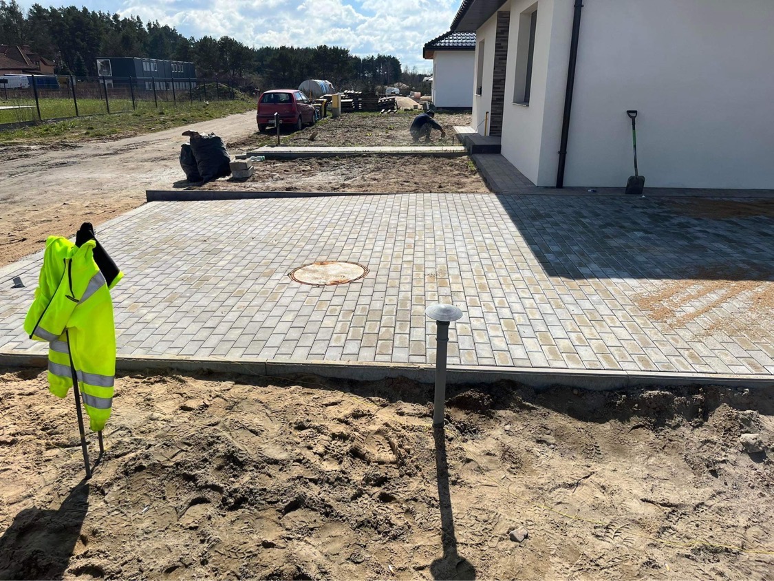 Ułożona kostka brukowa przed nowym domem, widoczna studzienka kanalizacyjna, lampa ogrodowa i odblaskowa kamizelka na kiju wbitym w piasek.
