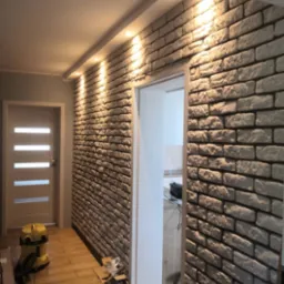 Korytarz z dekoracyjną ścianą z białej cegły, oświetloną punktowymi lampami sufitowymi, z widocznym odkurzaczem i otwartym opakowaniem materiałów budowlanych na podłodze.