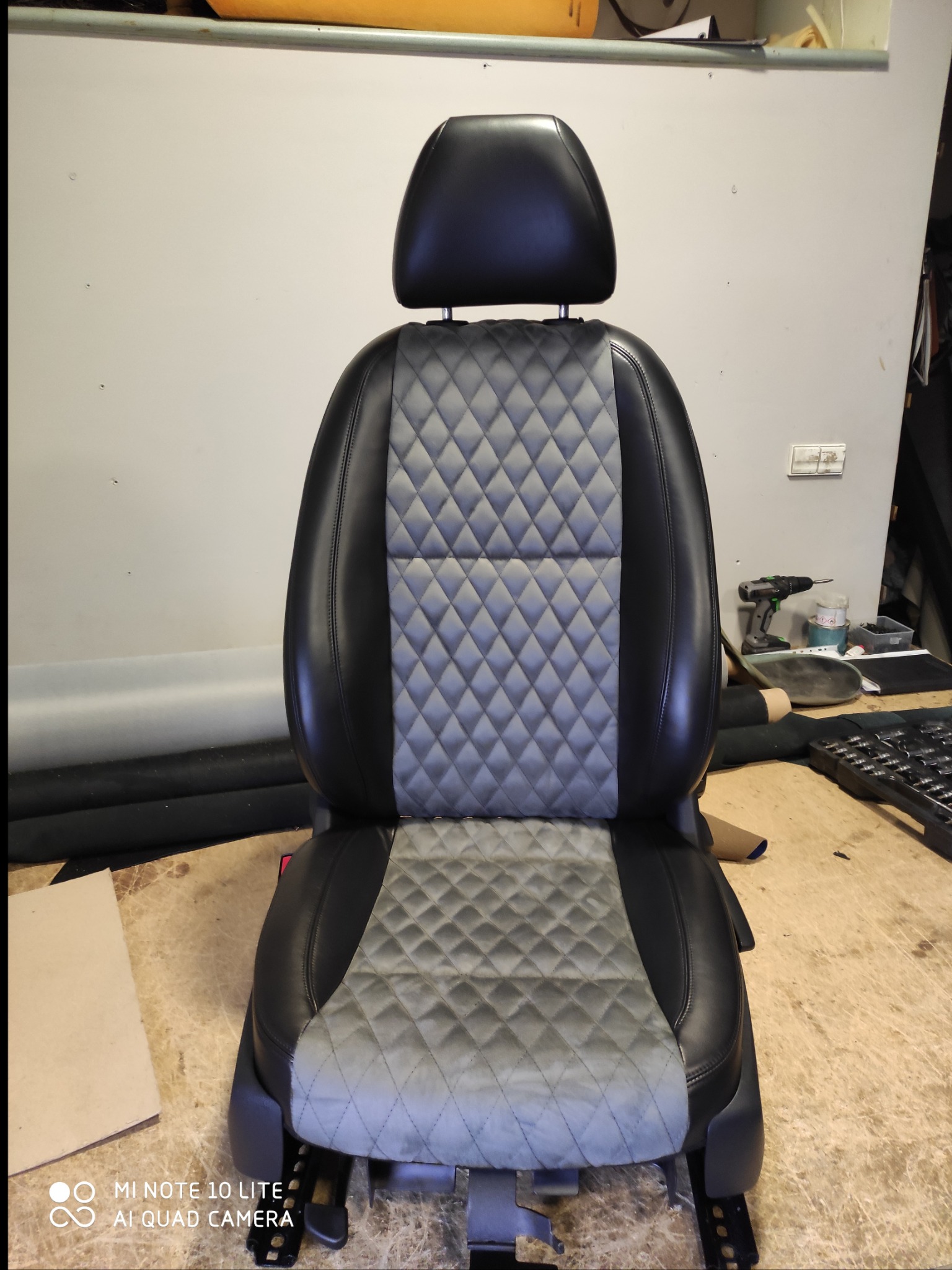 Fotel samochodowy po renowacji, czarna skóra po bokach, szare romby na siedzisku i oparciu, widoczne narzędzia i materiały tapicerskie w tle.