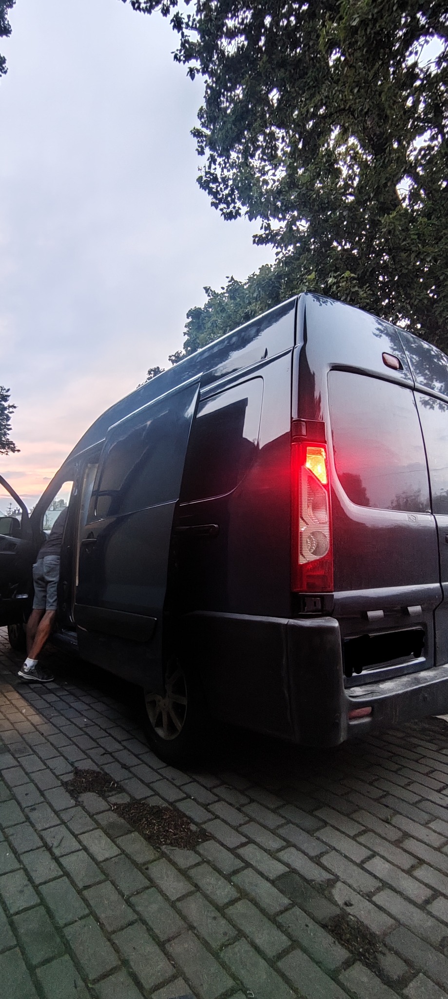 Ciemny bus z otwartymi drzwiami, częściowo widoczna sylwetka osoby wchodzącej, zapalona tylna lampa, kostka brukowa na ziemi, drzewa i zachodzące słońce w tle.