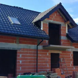 Niedokończony budynek z czerwonej cegły z zamontowanym czarnym dachem z dachówką, widoczne rynny, okno dachowe i drabina oparta o ścianę.