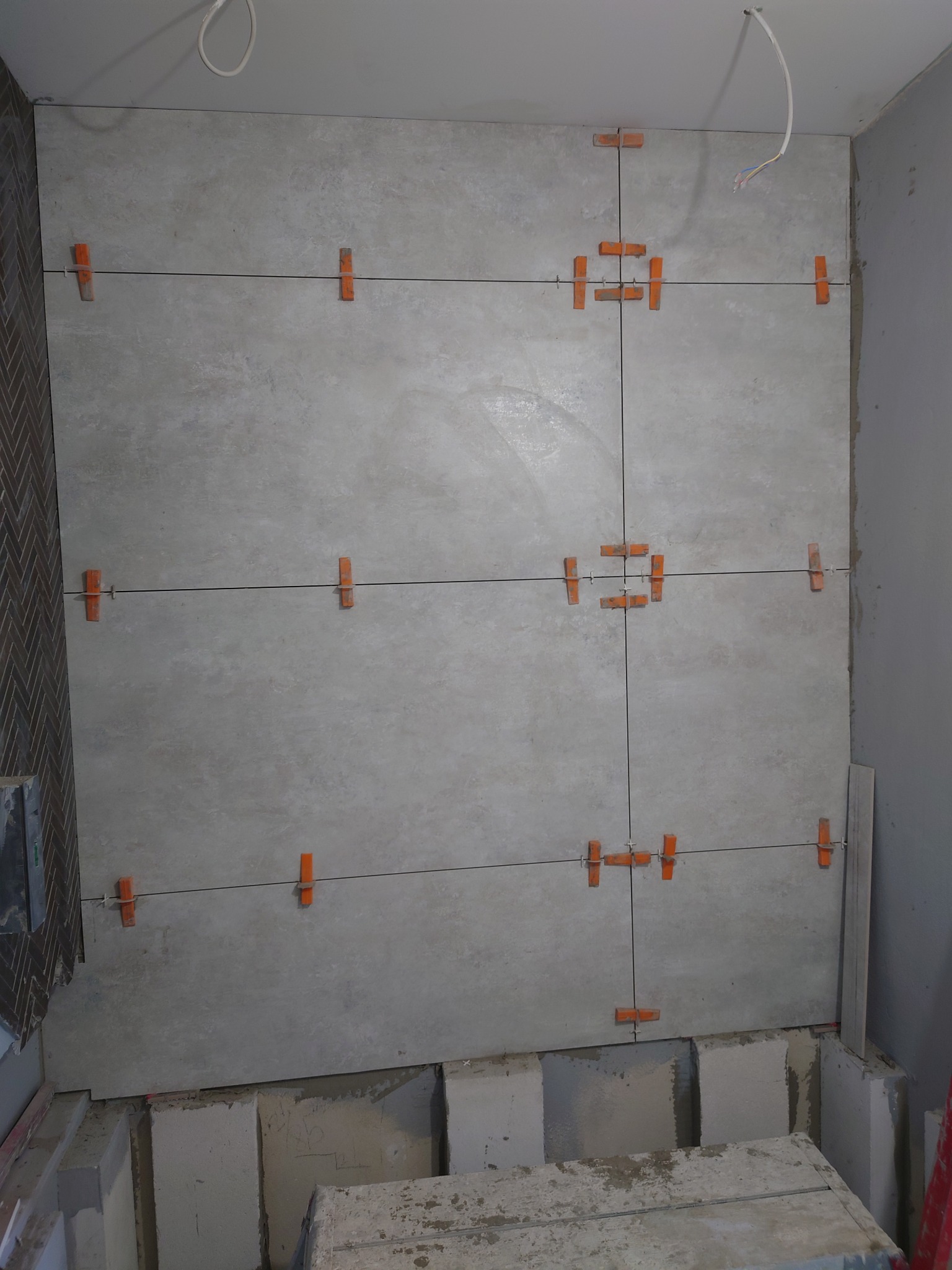 Układanie jasnoszarych płytek wielkoformatowych na ścianie z użyciem pomarańczowych krzyżyków dystansowych, widoczne fragmenty podłogi w trakcie prac remontowych.