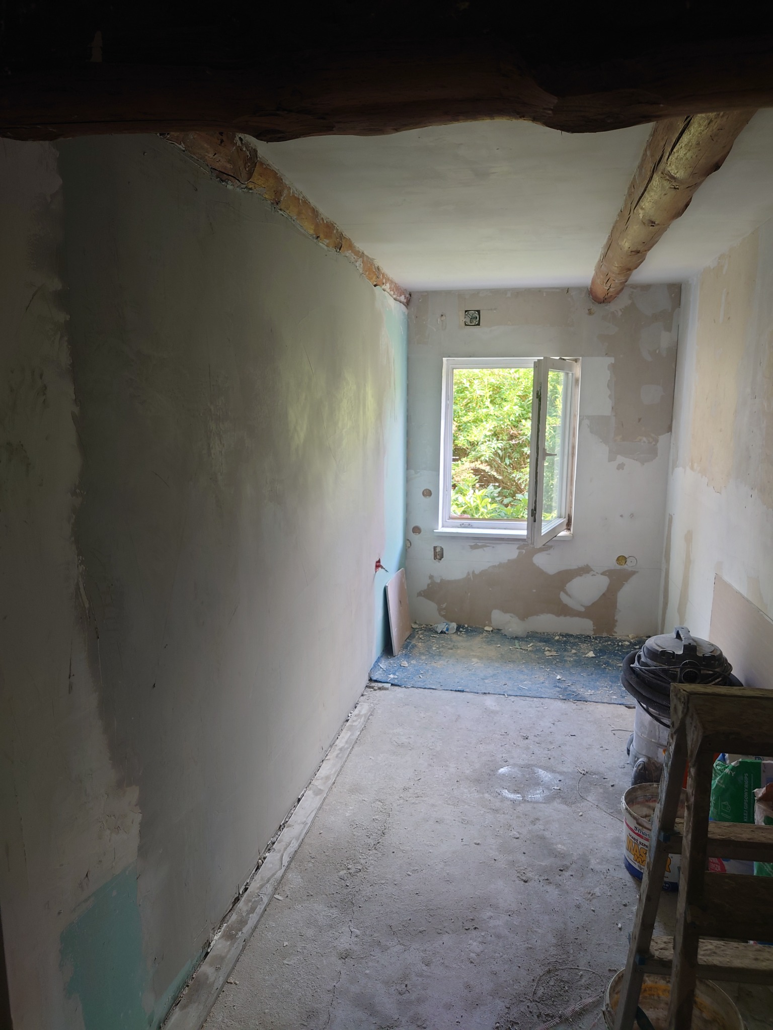 Pokój w trakcie remontu: ściana w szpachli, widoczne drewniane belki stropowe, okno z widokiem na zieleń, drabina oparta o ścianę, odkurzacz i wiaderka z farbą na podłodze.